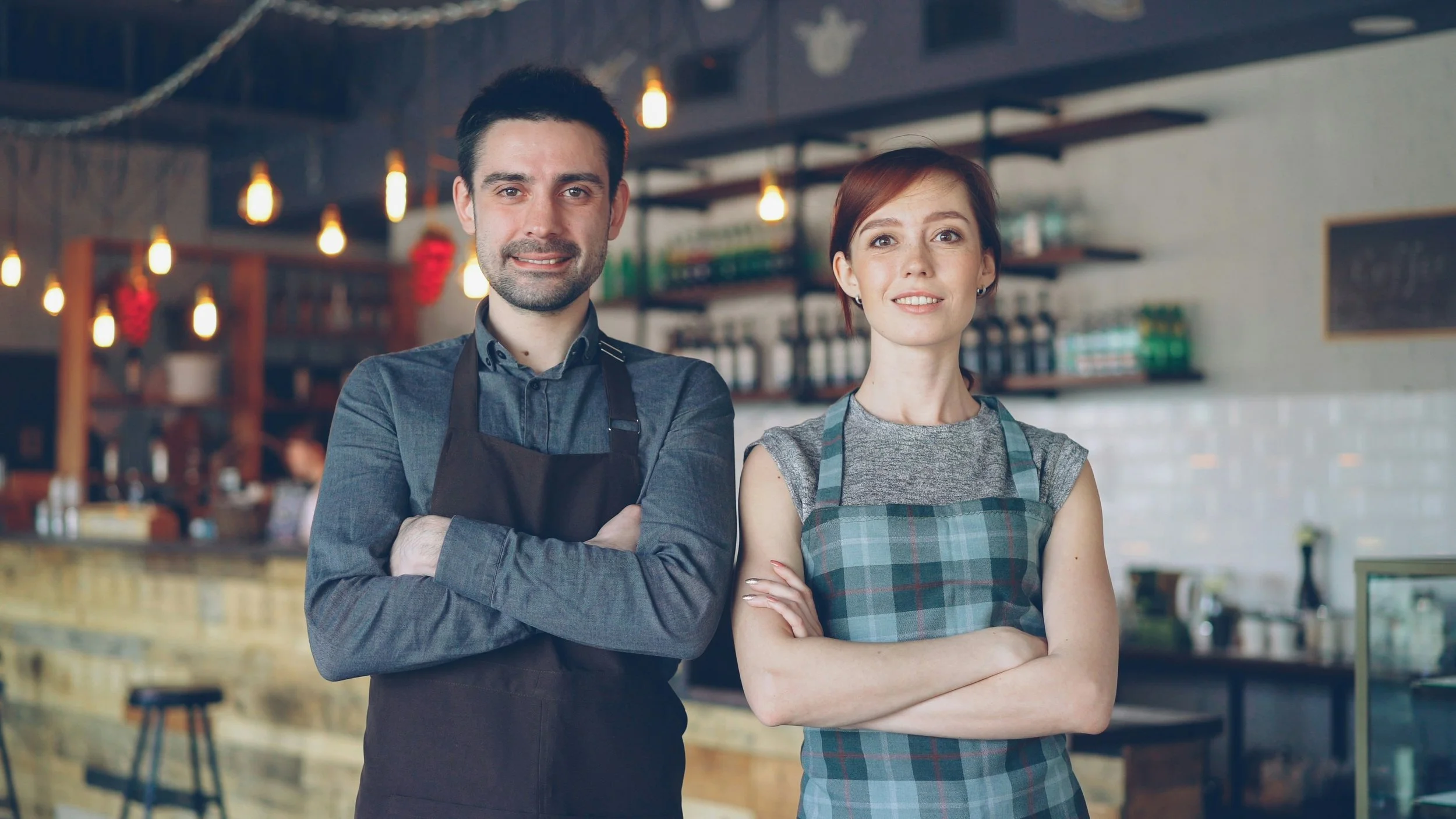 A man and a woman standing inside a cafe or restaurant, both wearing aprons with arms crossed and looking at the camera. The man has dark hair and a beard, and the woman has short reddish hair. The background shows a bar and shelves with bottles and 