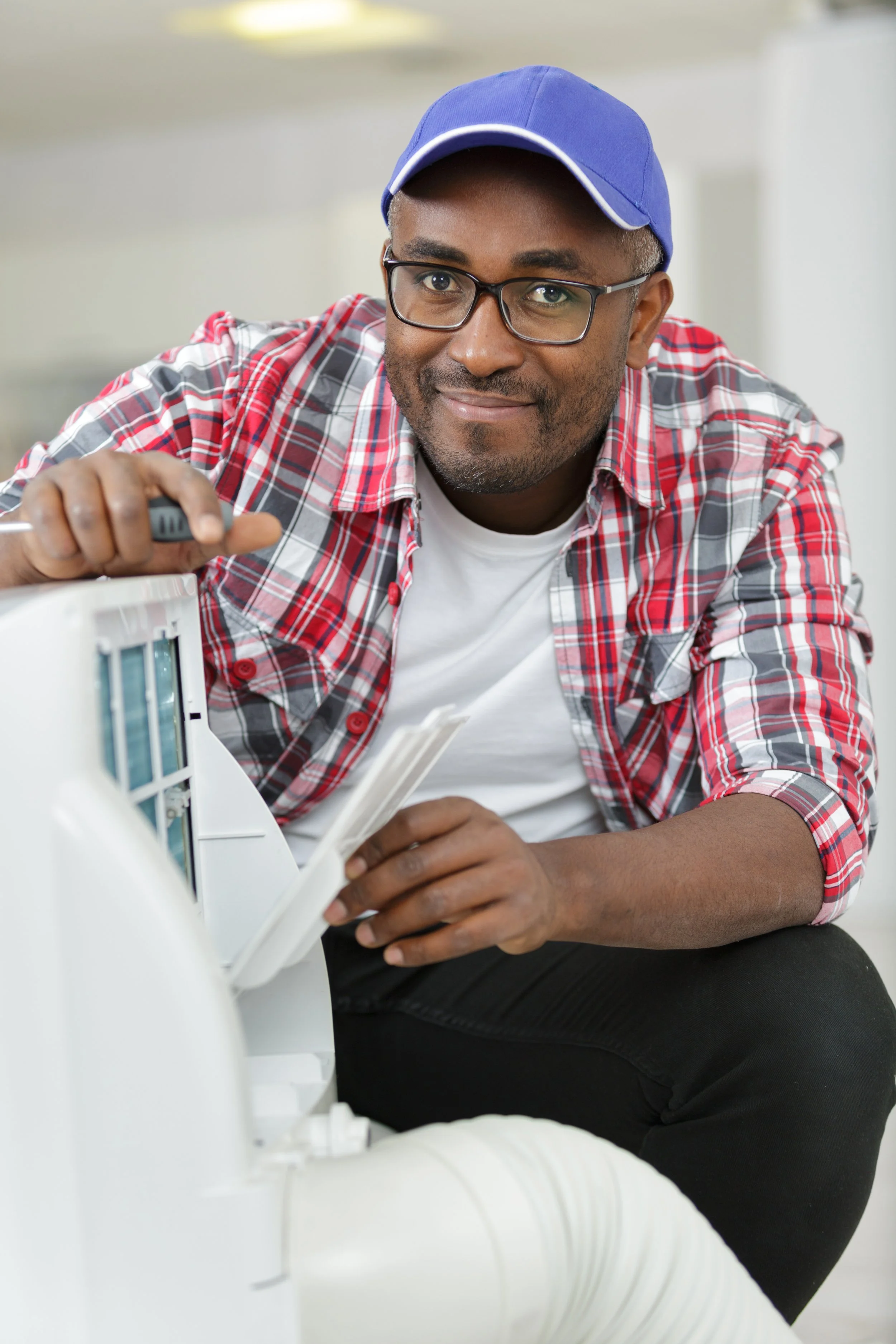 A smiling man wearing glasses, a blue cap, a red and gray plaid shirt, and a white T-shirt working on an appliance or machine in a well-lit room.