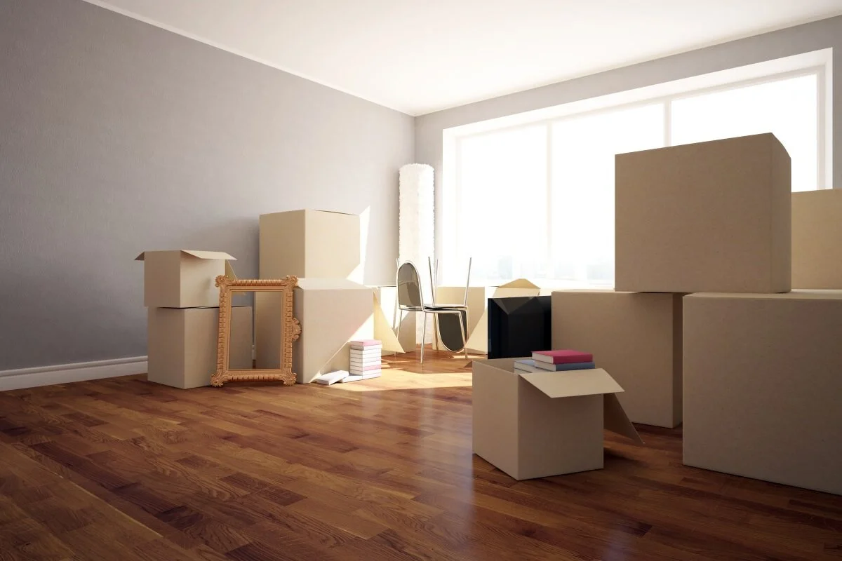 Room with cardboard boxes, a mirror frame, a chair, stacked books, and a large window with blinds.