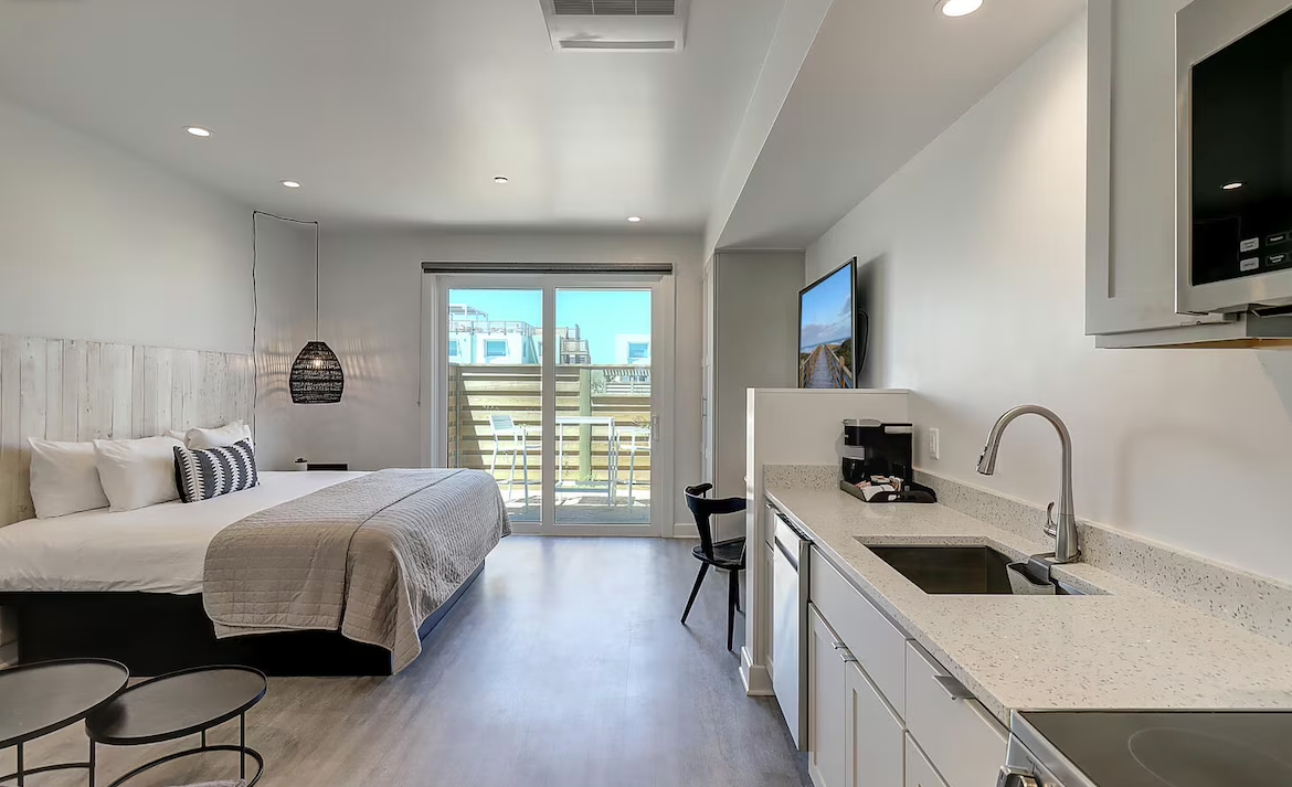 Studio apartment with a bed, small tables, a kitchenette, and a balcony with outdoor chairs.