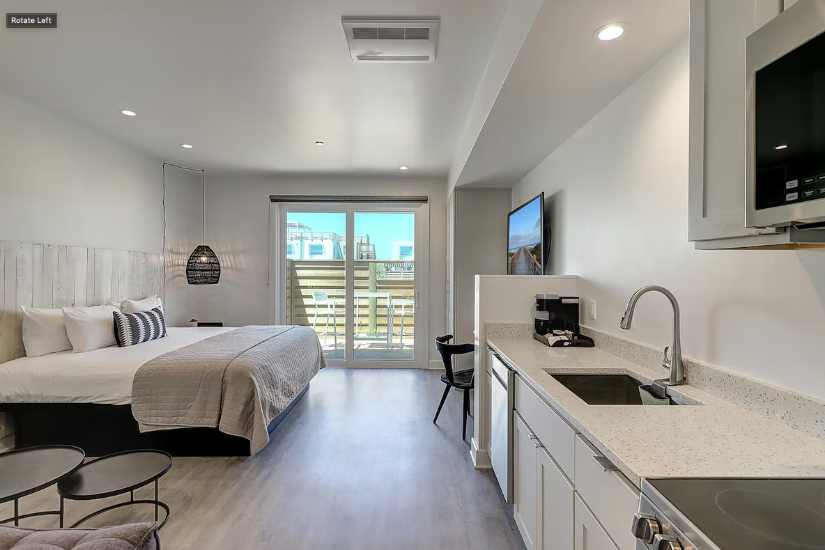 A modern studio apartment with a bed, small tables, a kitchenette with a sink, microwave, and stove, a flat-screen TV, and a balcony with a sliding glass door