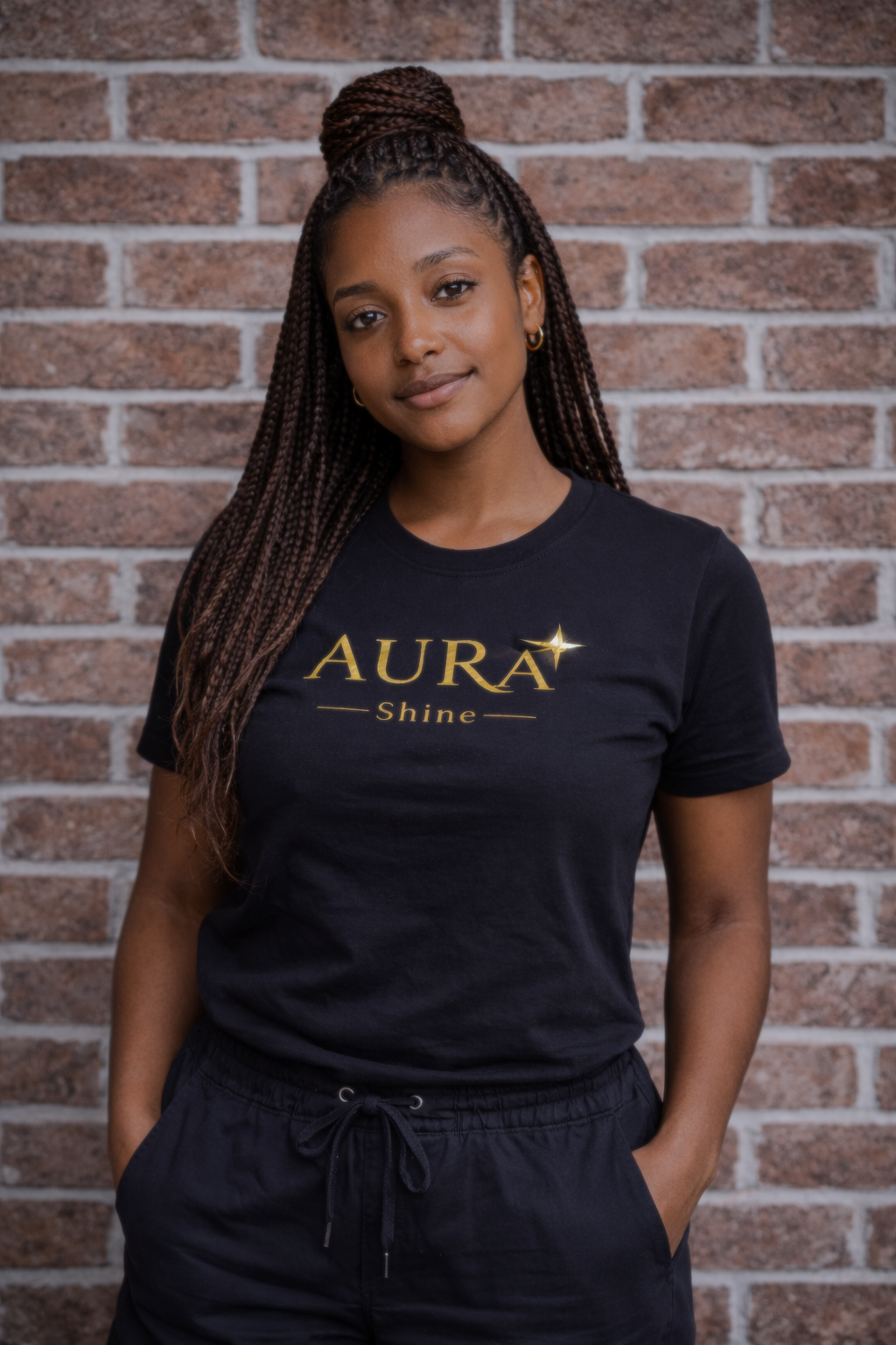 A young woman with braided hair standing against a brick wall, wearing a black t-shirt with the text 'AURA Shine' in gold and black pants with a drawstring.