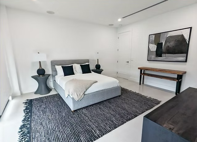 Modern bedroom with a white bed, black and white pillows, black bedside lamps, a black and white rug, a wooden bench, and a large abstract framed artwork on the wall.