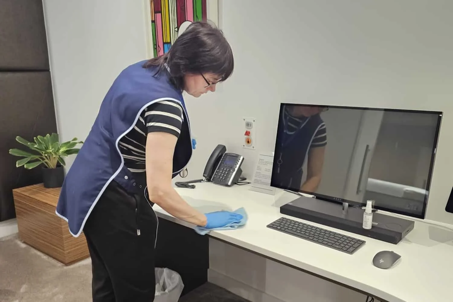 A woman cleaning a desk in an office, wearing a blue vest and gloves, with a plant, a computer monitor, keyboard, mouse, and telephone on the desk.