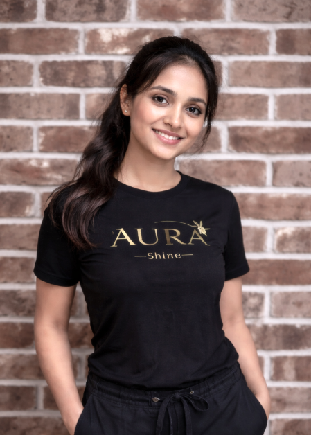 A young woman with long dark hair smiling, wearing a black t-shirt with the words "AURA Shine" printed on it, standing in front of a brick wall.