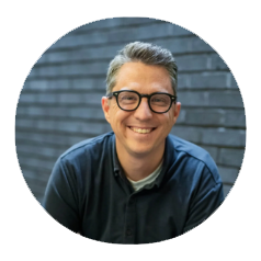 A smiling man with glasses and gray hair, wearing a dark blue shirt, sitting in front of a gray brick wall.