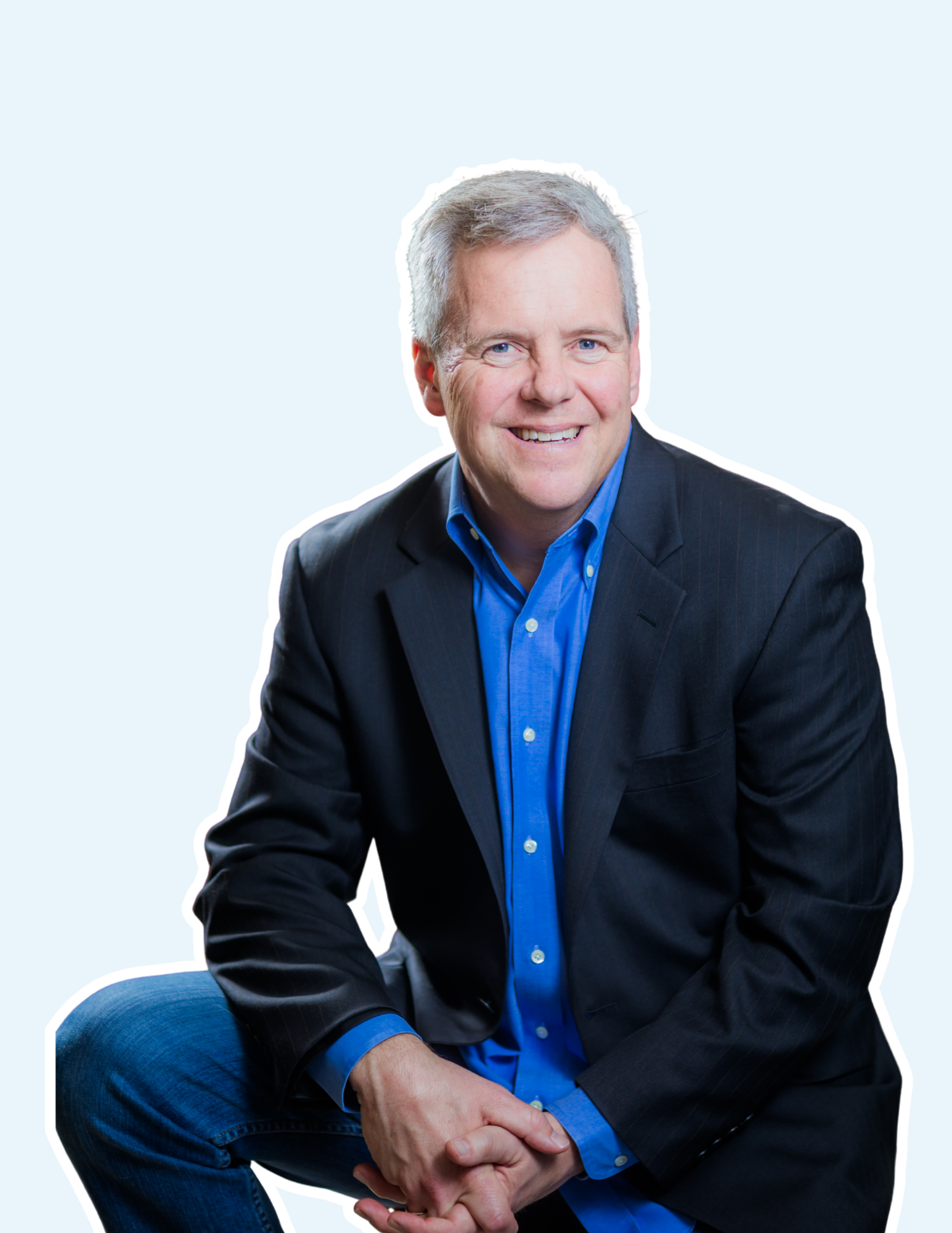 A man with short gray hair, wearing a dark suit jacket and blue shirt, smiling and squatting with hands clasped in front of him against a light background.