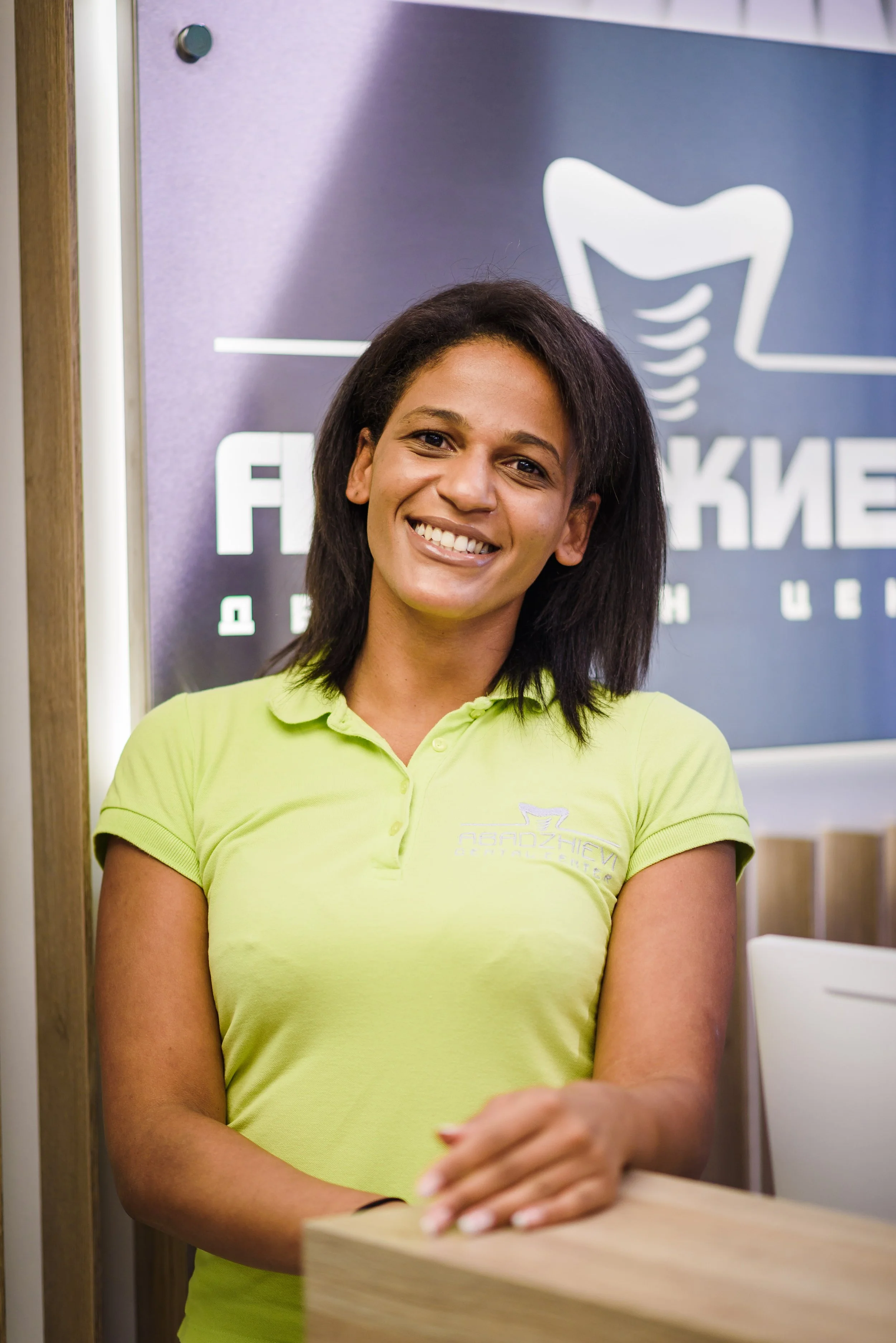 Young woman with long brown hair smiling and sitting on a counter in front of a sign that reads 'iDENTITY' with the tagline 'Your smile is your identity' in an orthodontics or dental office.