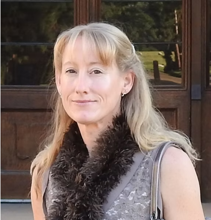 A woman with blonde hair standing outdoors in front of a wooden building with glass windows.