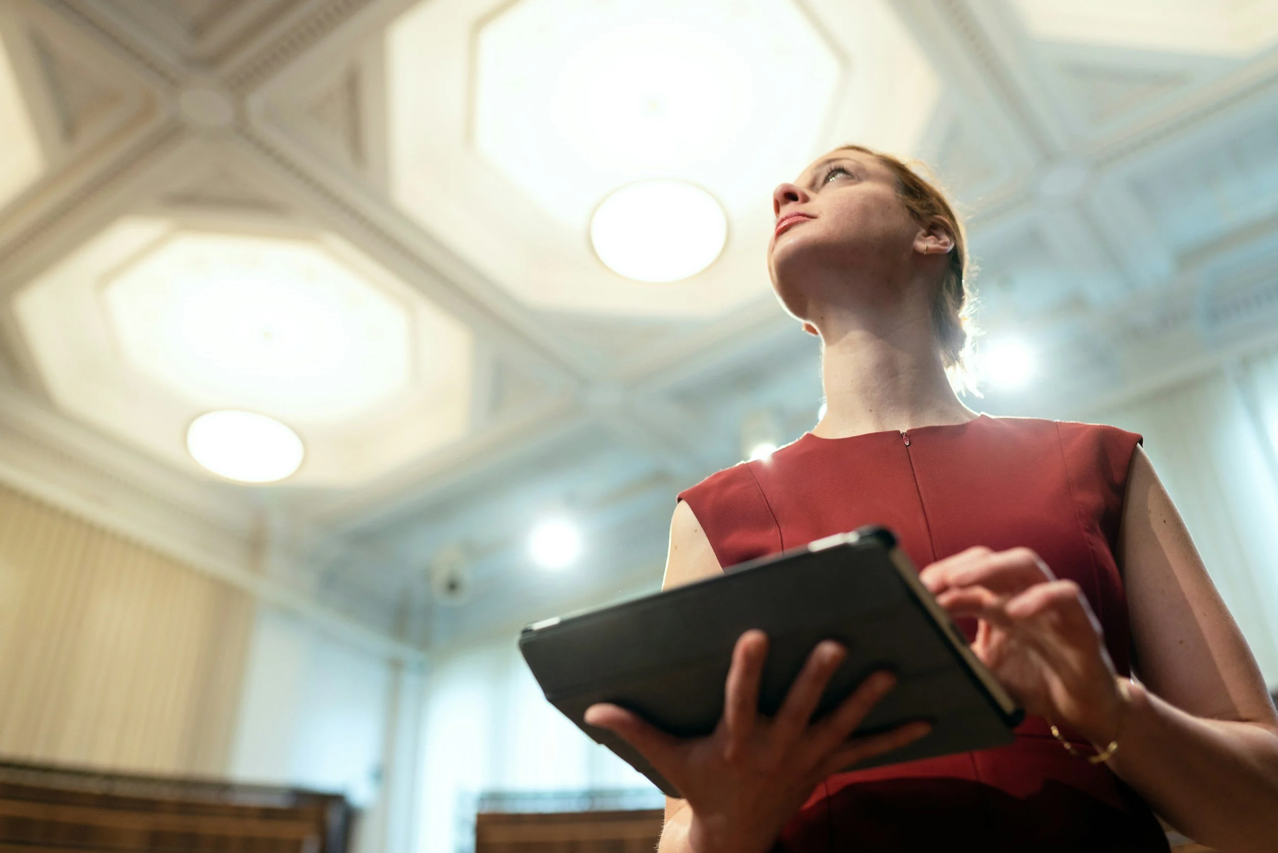 Una donna con capelli rossi, indossa un vestito rosso e tiene in mano un tablet, si trova in un ambiente elegante con soffitto decorato e illuminazione diffusa.