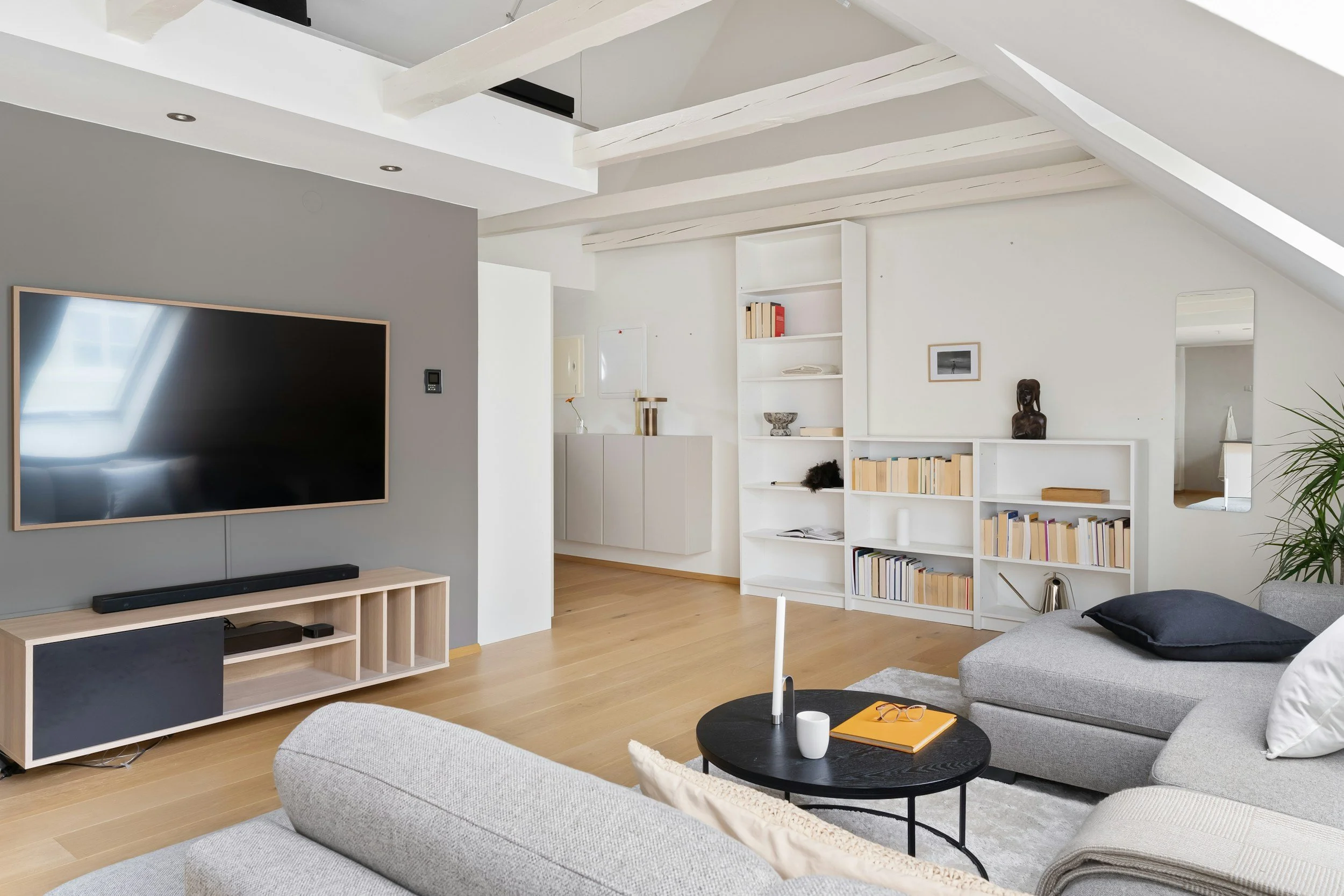 Interno di un soggiorno con divano grigio chiaro, tavolino nero con oggetti e sedie, pareti bianche, libreria bianca e televisore montato su parete grigia.
