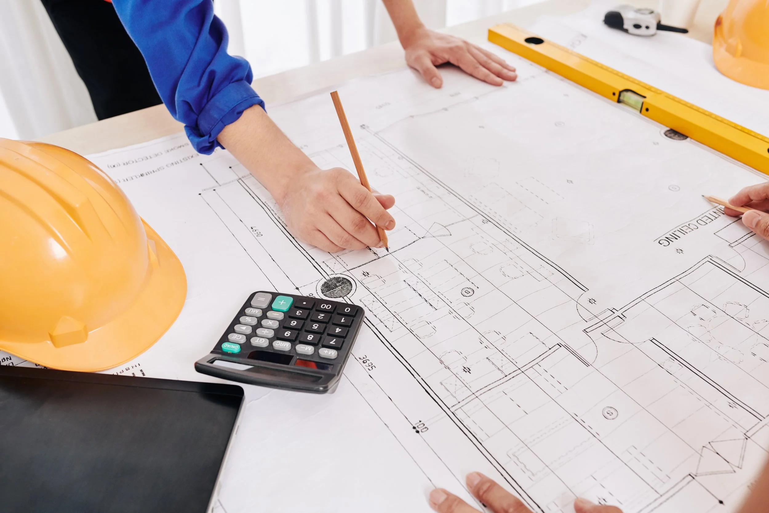 Persona che lavora su un progetto di architettura con piante tecniche su un tavolo, con strumenti da disegno, elmetto di sicurezza, calcolatrice e livello laser.
