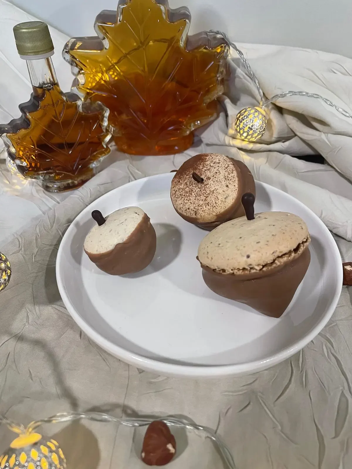 Macarons au chocolat posés sur une assiette blanche, avec deux bouteilles de forme de feuilles d'érable en verre en arrière-plan, décorés avec des petites lumières décoratives.
