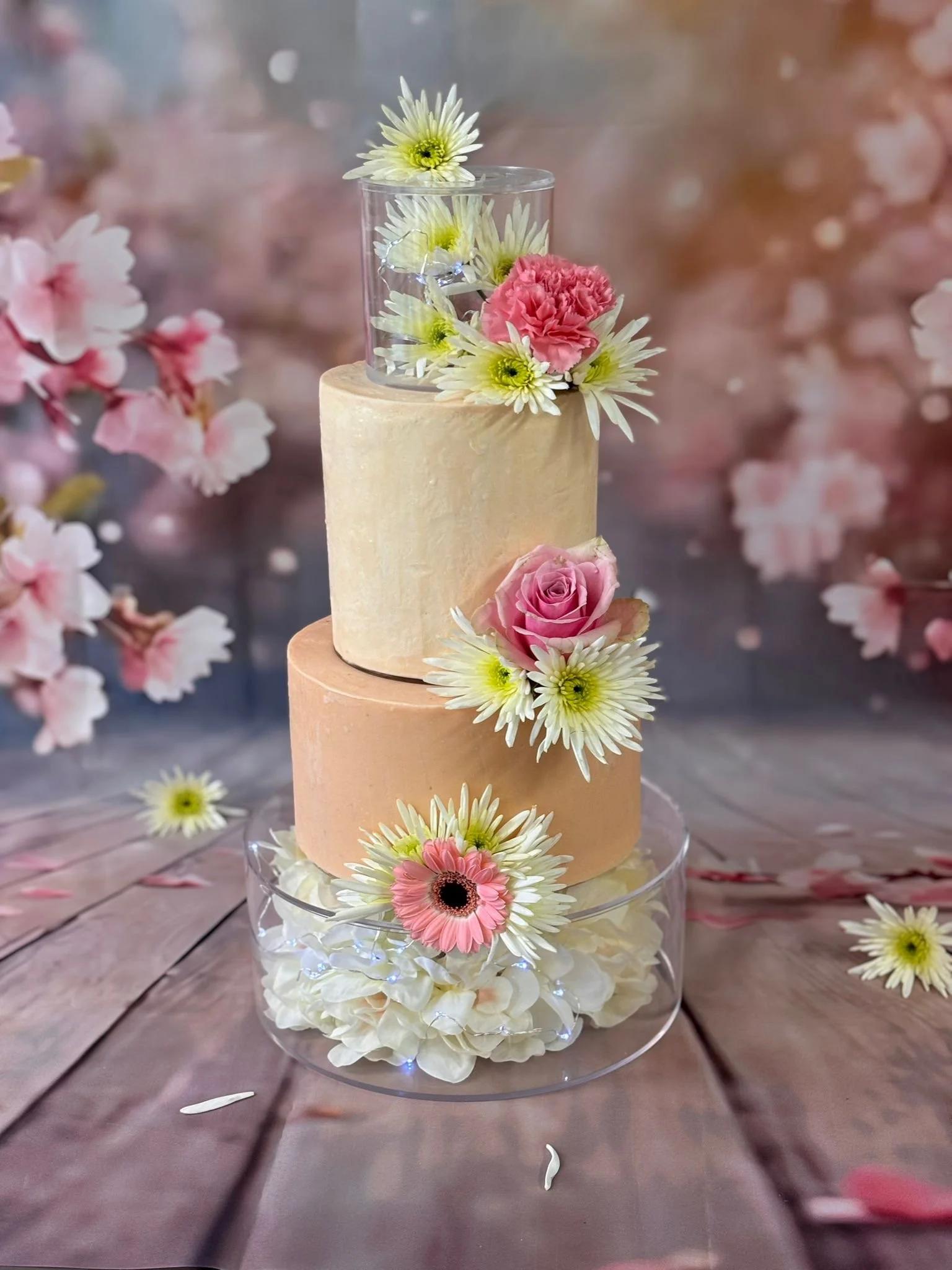 Gâteau à trois niveaux avec décor floral comprenant des roses roses, des marguerites blanches et des hortensias blancs, entouré de fleurs sur un fond flou de fleurs.