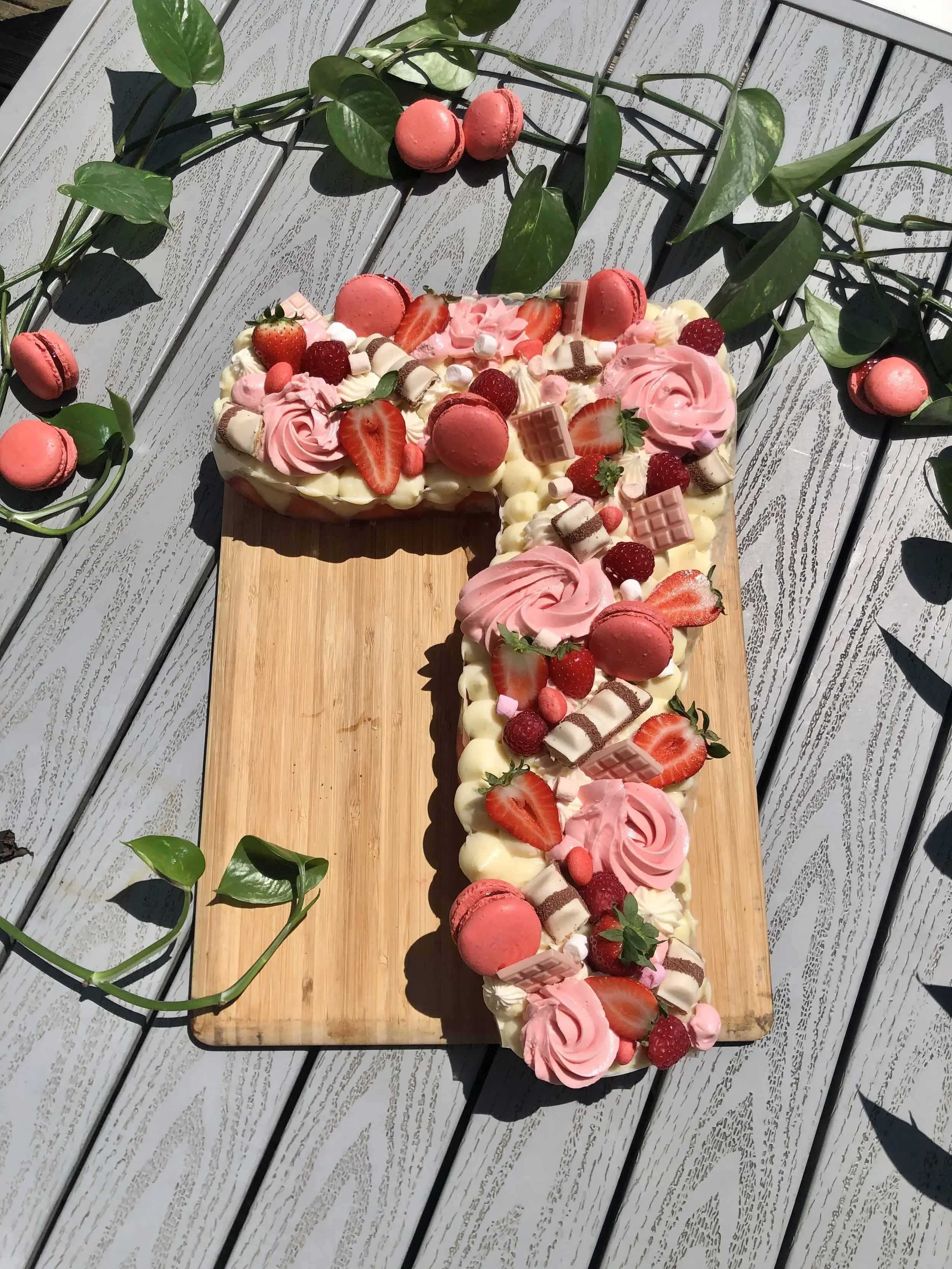 Gâteau en forme de lettre T décoré avec des fraises, macarons roses, biscuits, chocolat, et crème rose, posé sur une planche en bois avec des brins de feuillage vert autour.