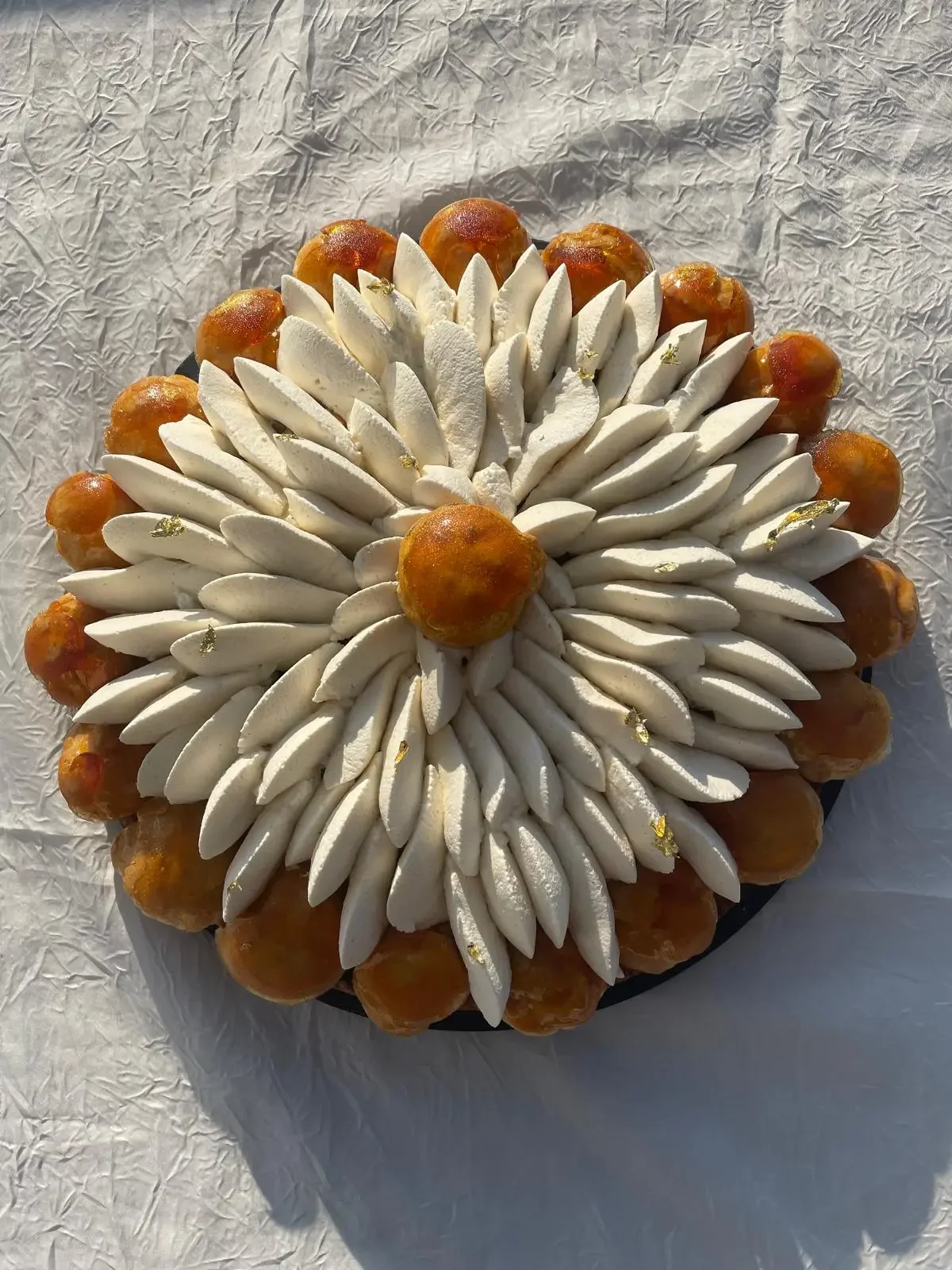 Gâteau décoratif orné de meringues blanches et de fruits orange, agrémenté de feuilles d'or comestibles, posé sur un fond blanc froissé.