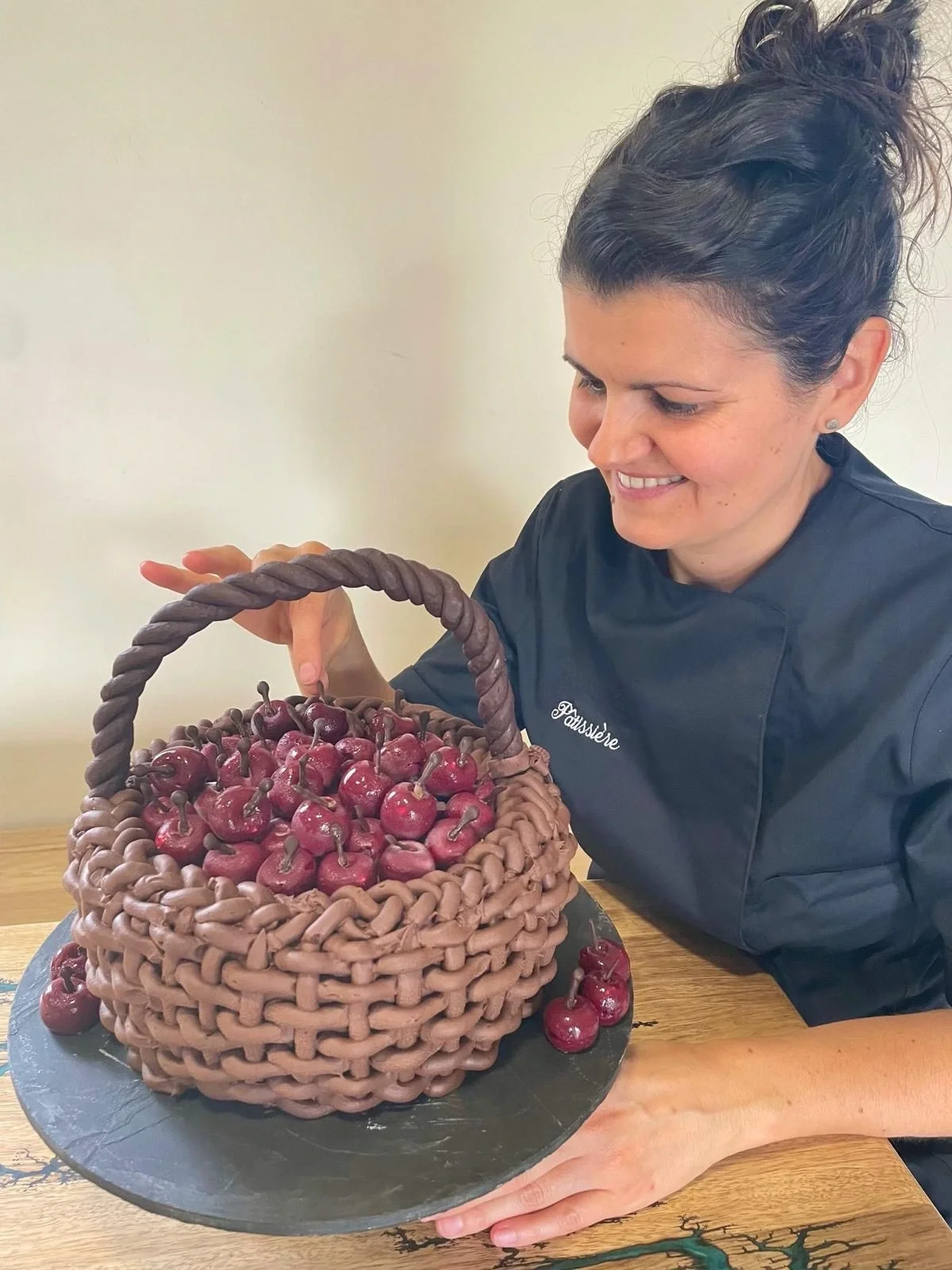 Une femme souriante en tenue de chef regarde un gâteau en forme de panier en chocolat rempli de cerises en chocolat.