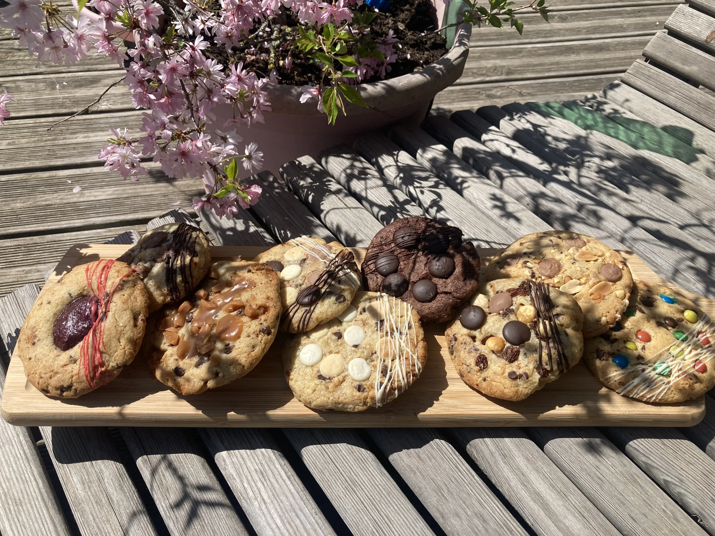 Un plateau de cookies décorés avec différentes garnitures, placé sur une table en bois avec une plante de fleurs roses en arrière-plan.