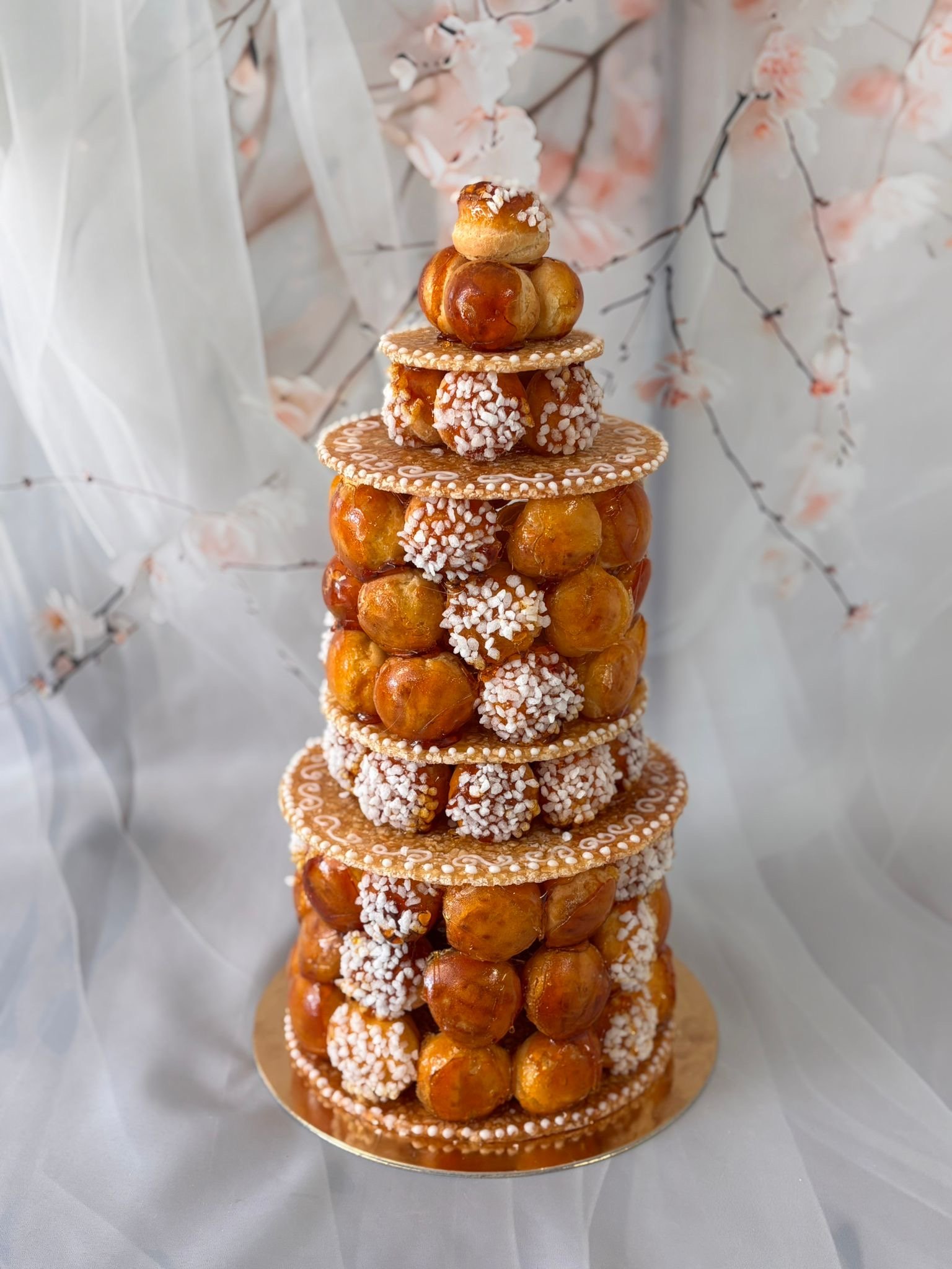 Gâteau décoratif pyramidal composé de choux, biscuits et éclats de sucre, avec un décor floral en arrière-plan.