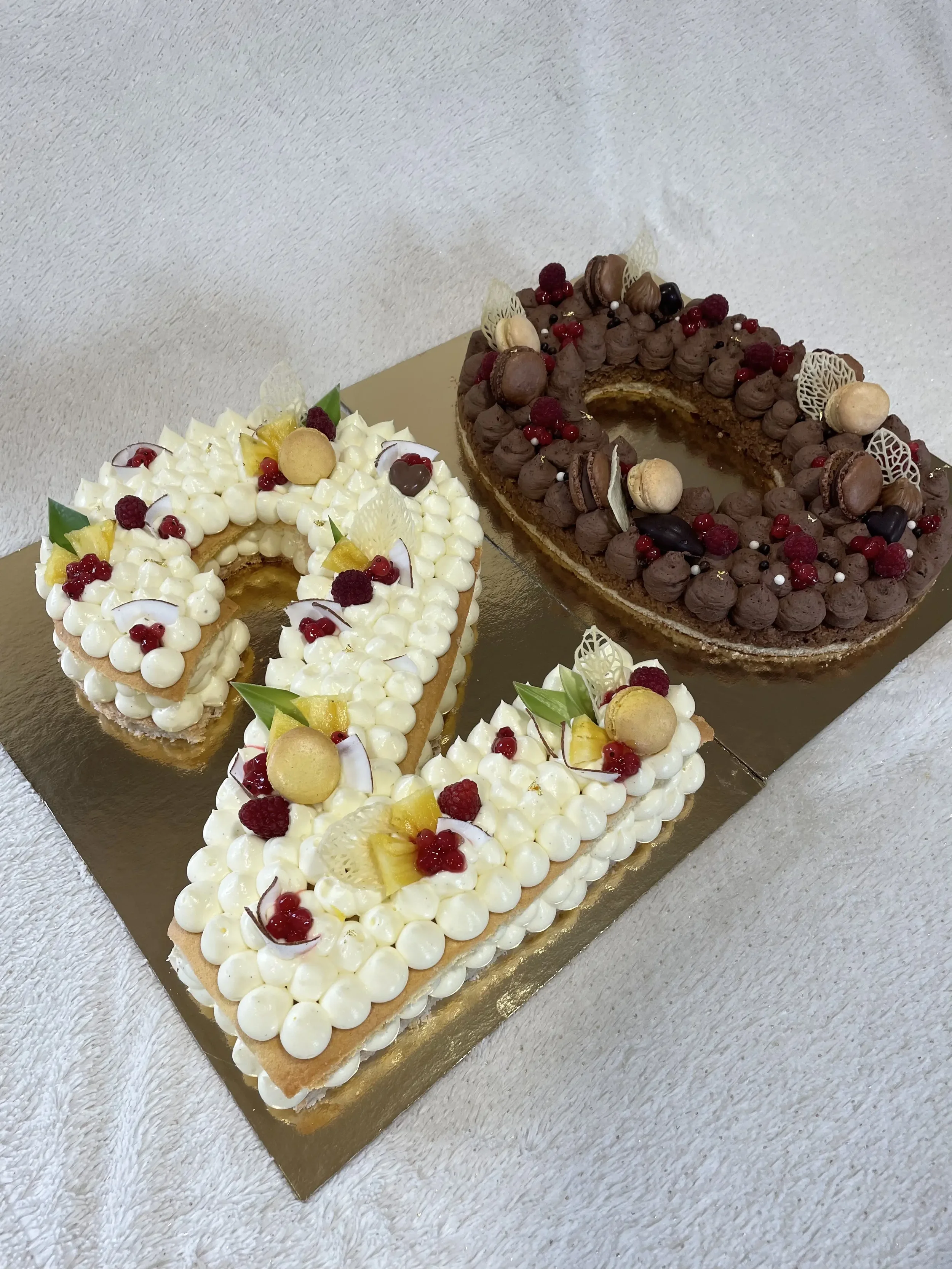 Deux gâteaux de forme rectangulaire décorés de crème blanche, fruits et chocolats, et un gâteau en forme de chiffre quatre décoré de crème, fruits et chocolats, sur une surface dorée.