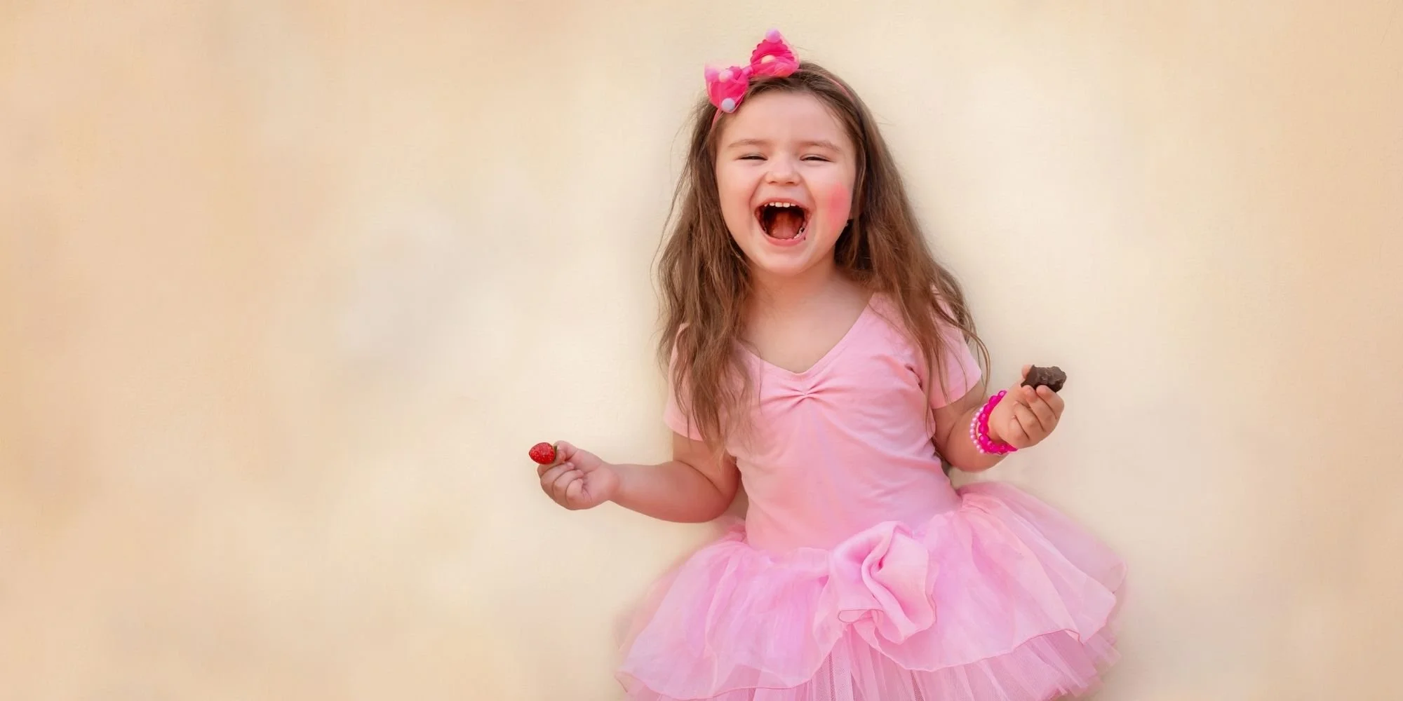 A young girl wearing a pink dress with a bow in her hair and pink bracelets, smiling and laughing while holding a chocolate treat and a strawberry.