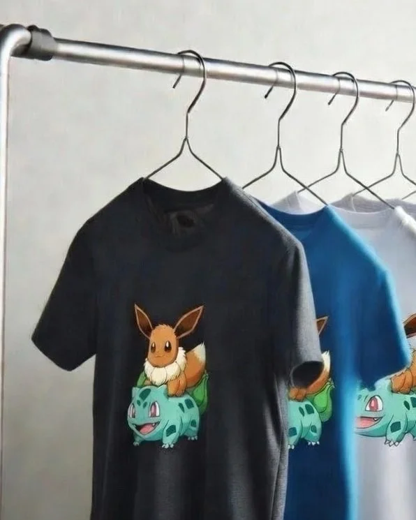 Three T-shirts hanging on a metal rod featuring a Cute Brown Fox Riding Green Plant Dinosaur, with the T-shirts in black, blue, and white.