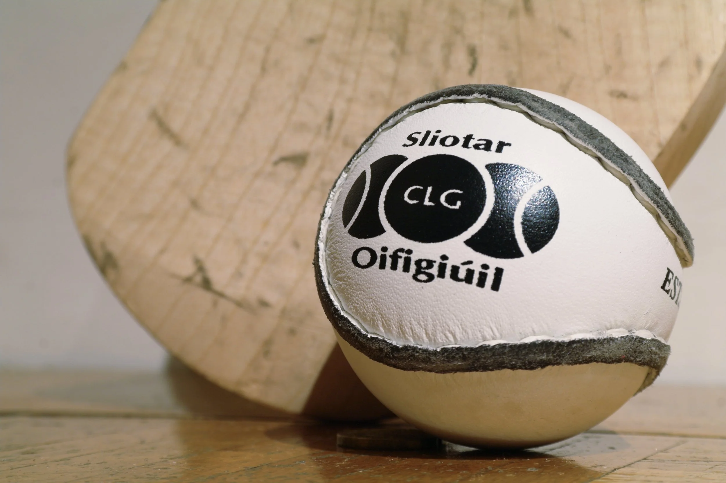 Close-up of a worn-out white ball with black text that reads 'Sliotar Oifigiúil' and 'CLC'. The ball is placed on a wooden surface with a wooden background.