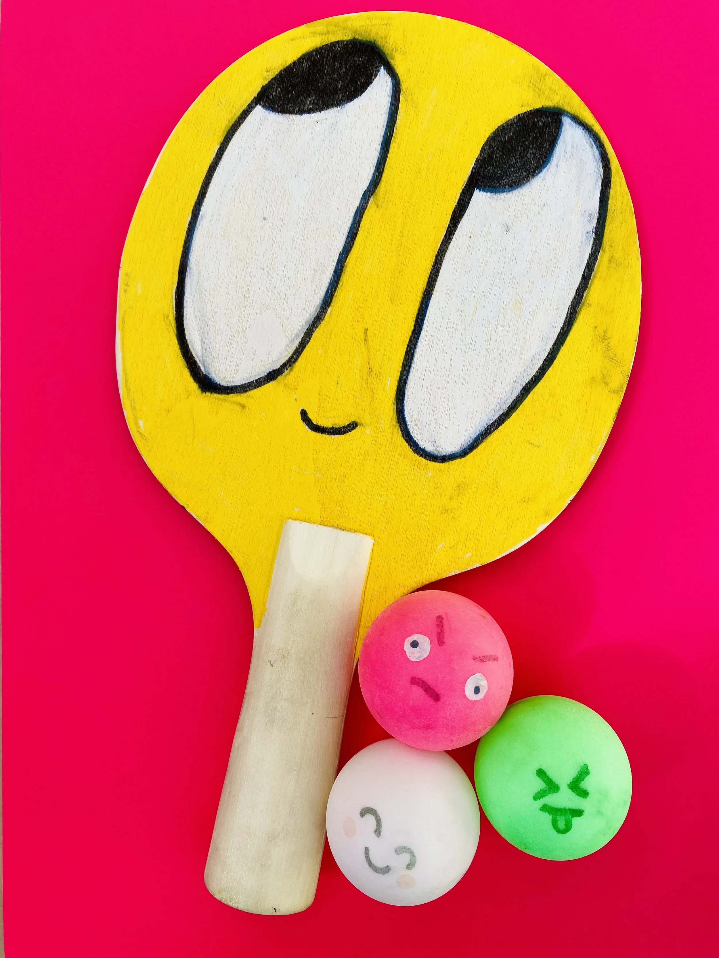 A wooden ping pong paddle with a painted emoji face, surrounded by four foam balls with various facial expressions, on a bright pink background.