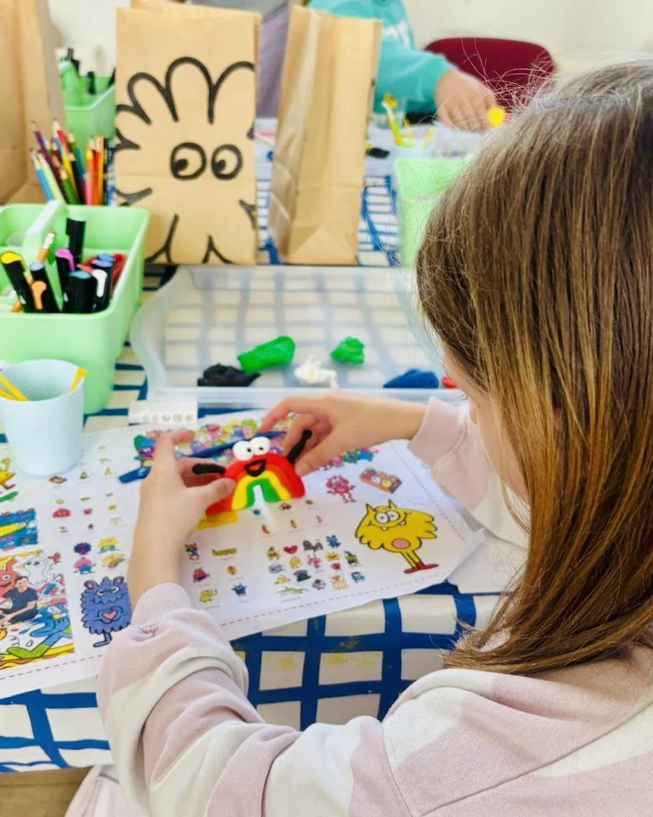 A child with brown hair working on a craft project that features a colorful, creature-like figure with eyes, a smile, and bug-like antennae. The table is covered with sheets of stickers, and in the background, a paper bag with a cartoon face and other craft supplies are visible.