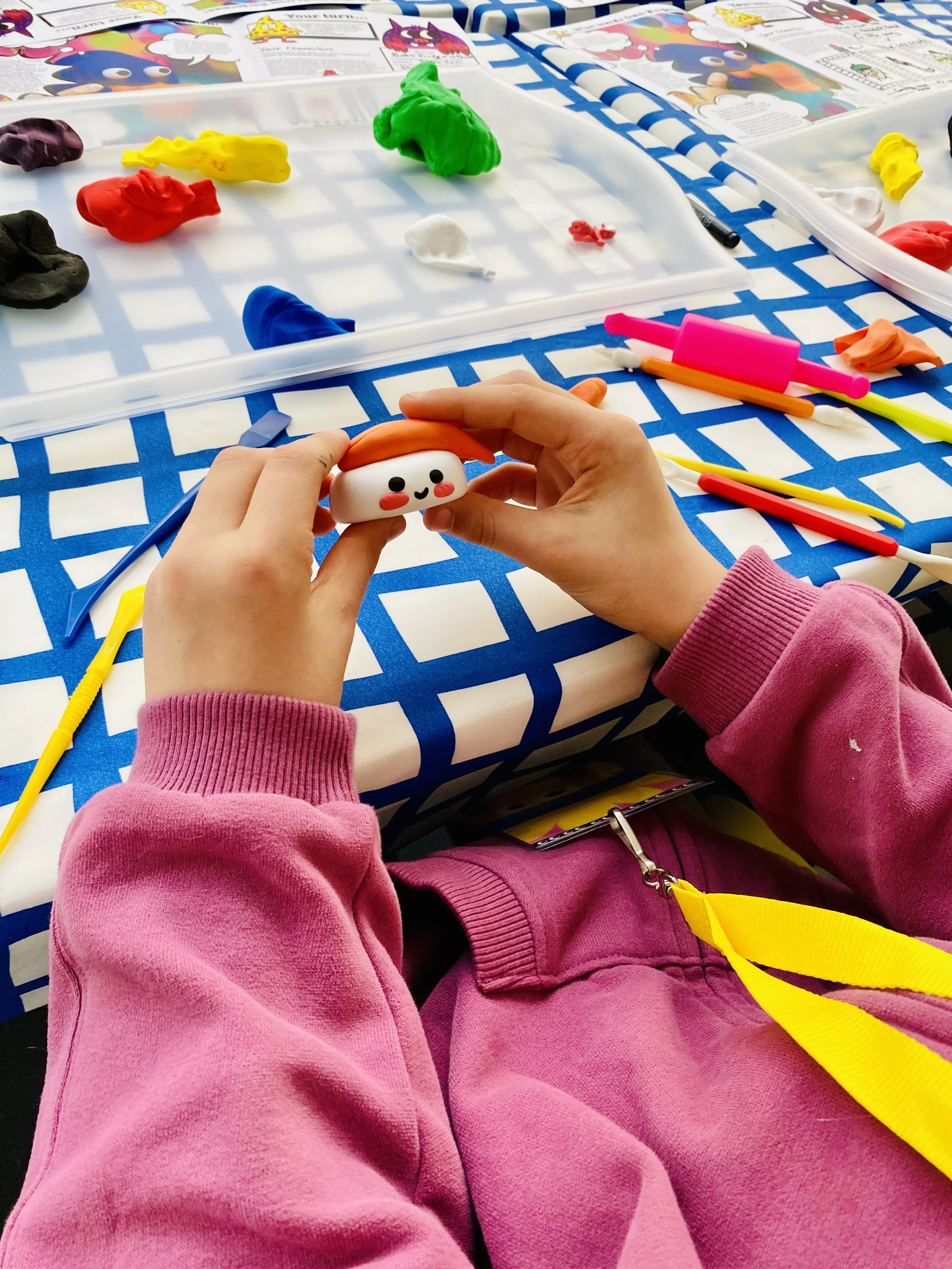 Person wearing pink sweatshirt sitting at table with colorful playdough and modeling tools, holding a small, smiling, cartoon-like food figure.