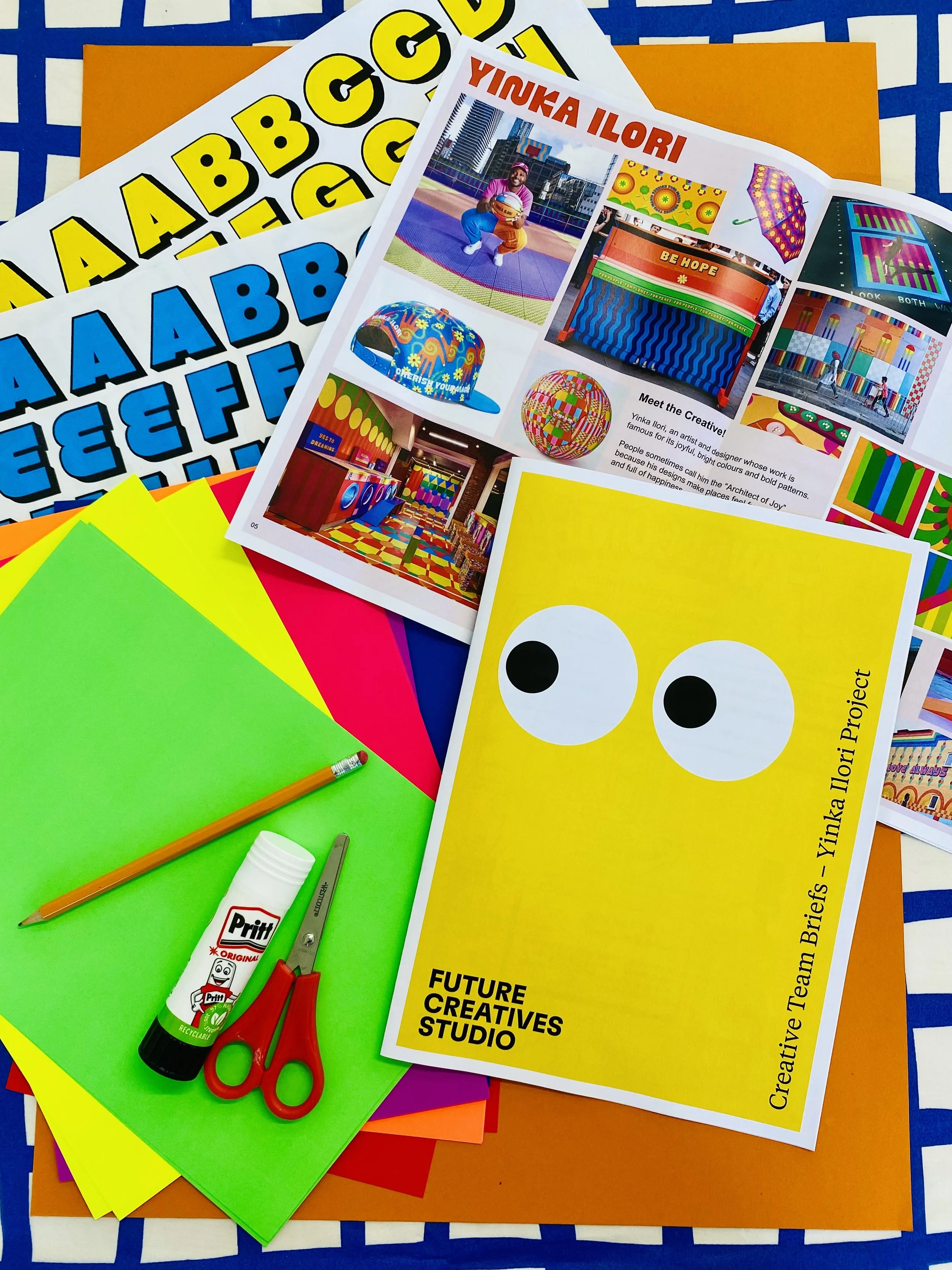 Colorful paper sheets with scissors, glue stick, and a pencil arranged on top of a surface, along with project flyers featuring vibrant images of an artist and art installations.