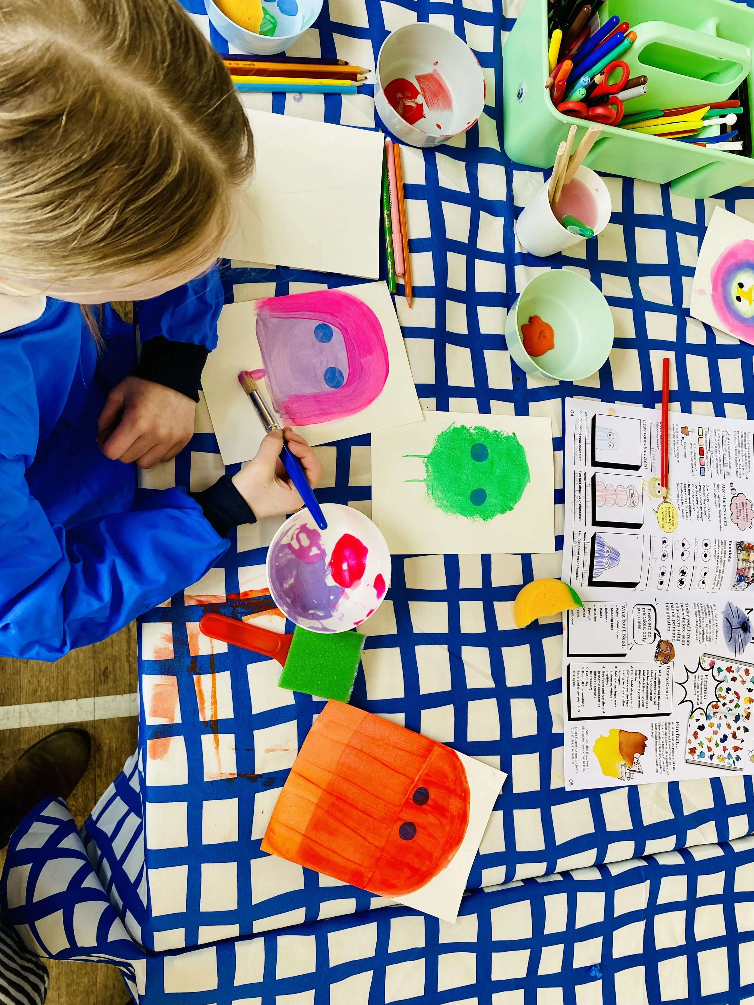 A child painting colorful artwork at a table covered with a blue and white checkered tablecloth. The table has various art supplies, including paint, brushes, markers, scissors, and paper with painted faces and designs.