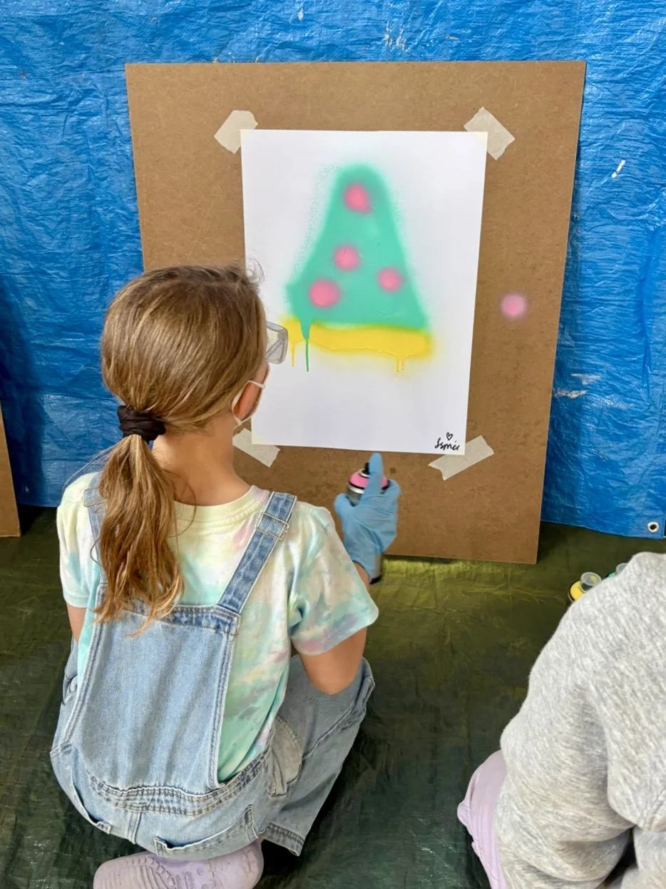 A girl with long hair tied in a ponytail, wearing glasses, a tie-dye t-shirt, and denim overalls, is spray painting a Christmas tree on a white paper attached to a brown board with masking tape. The background includes a blue tarp and a person partially visible wearing a gray hoodie.