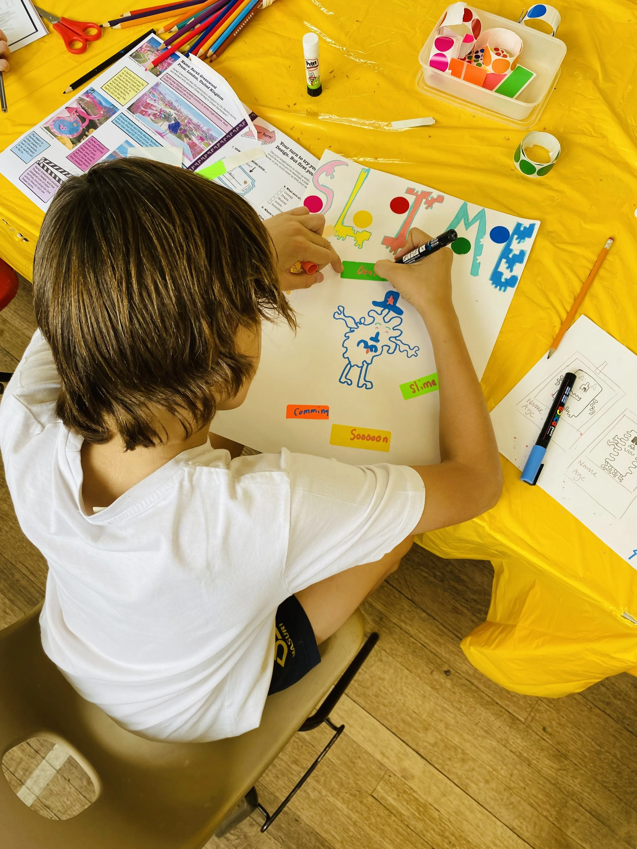 A person with shoulder-length brown hair drawing a colorful poster at a table covered with a yellow tablecloth, surrounded by art supplies like markers, tape, and paper.