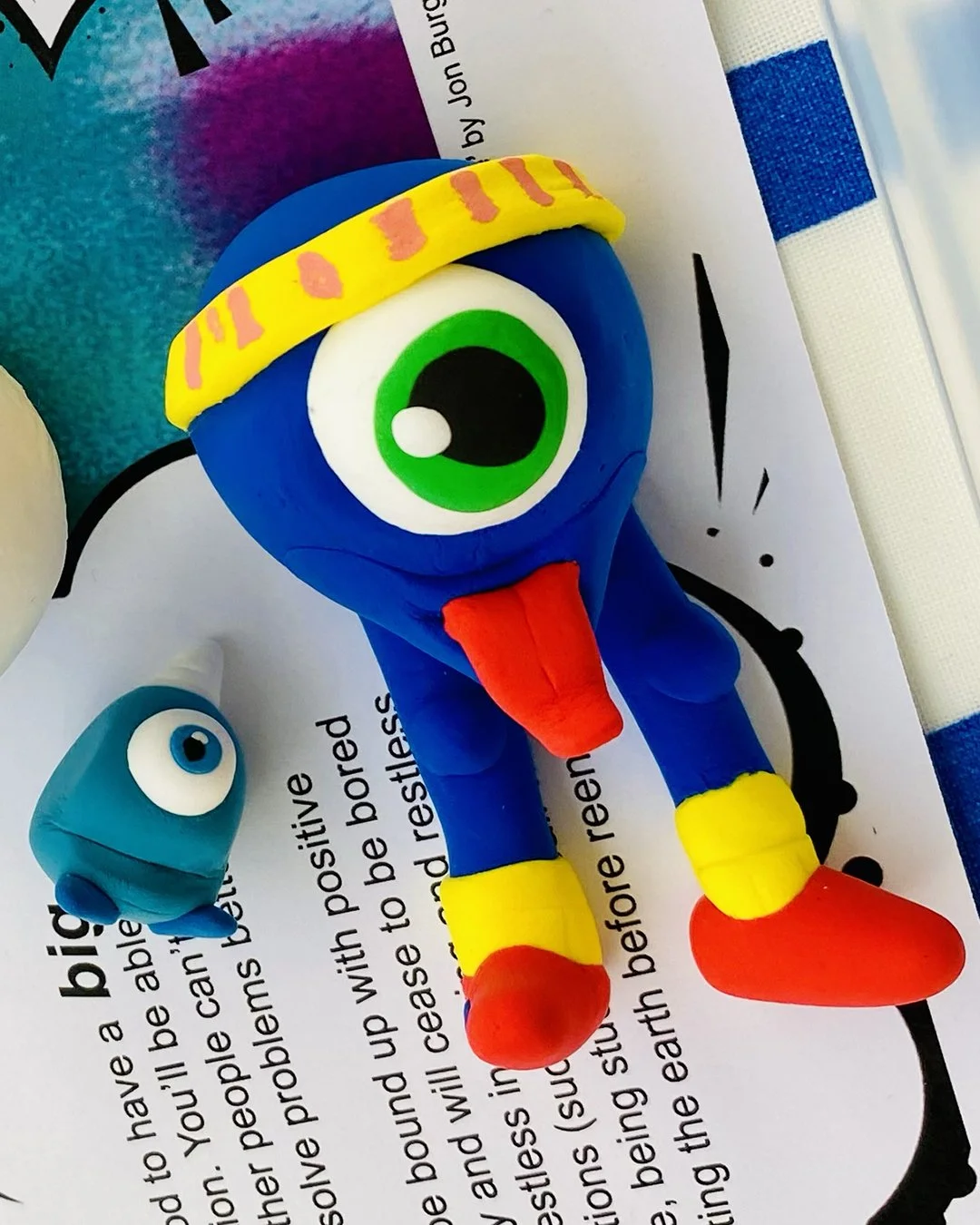 Colorful clay sculpture of a blue cartoon character with one large green eye, wearing a yellow hat with pink markings, yellow shoes with red tips, and a red tongue sticking out. The sculpture is placed on a printed sheet of paper with text.