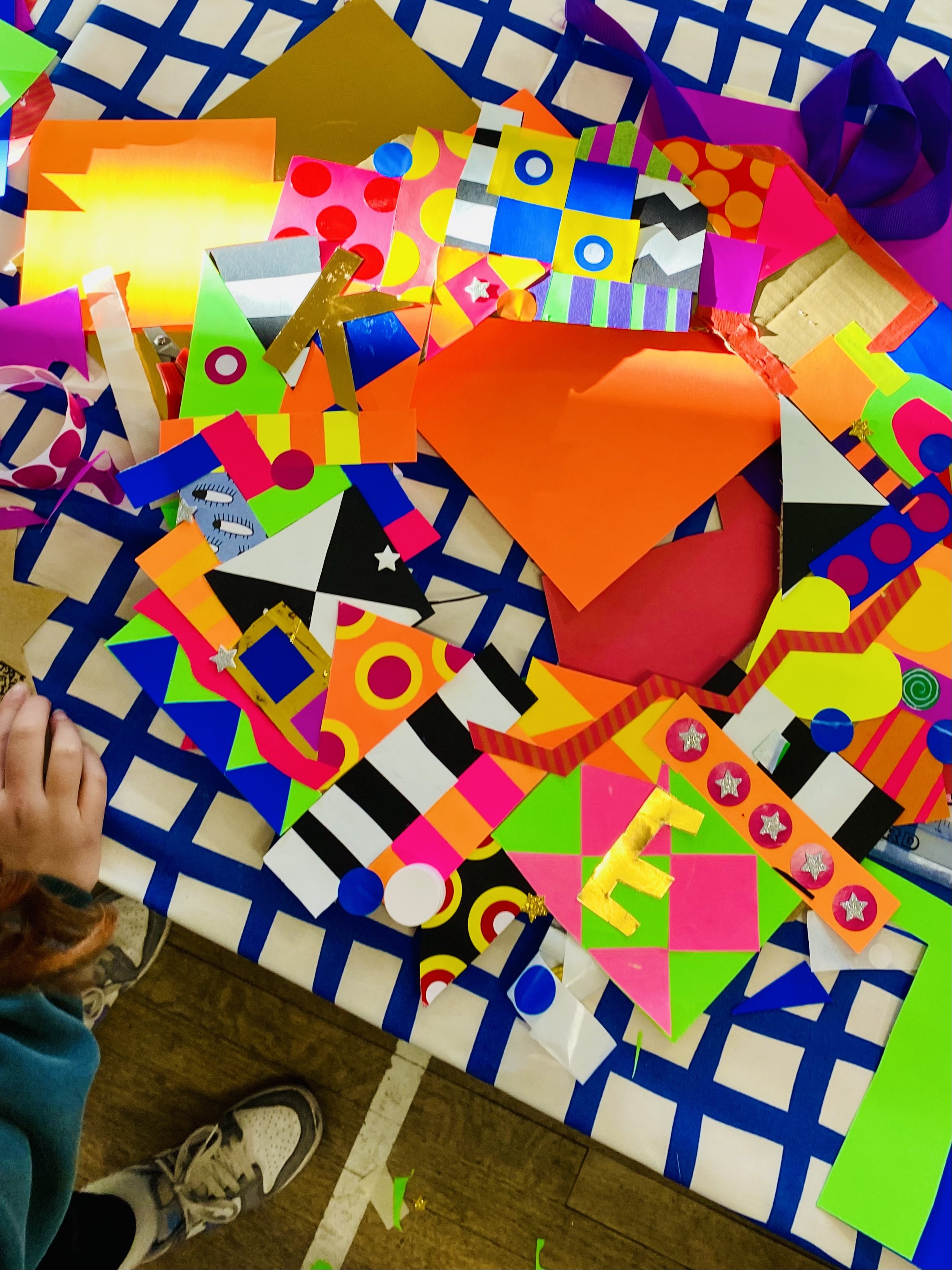 Colourful cut cards and stickers stuck together to make a collage wreath on a blue and white checkered tablecloth, with children's shoes and a child's hand visible in the corner.