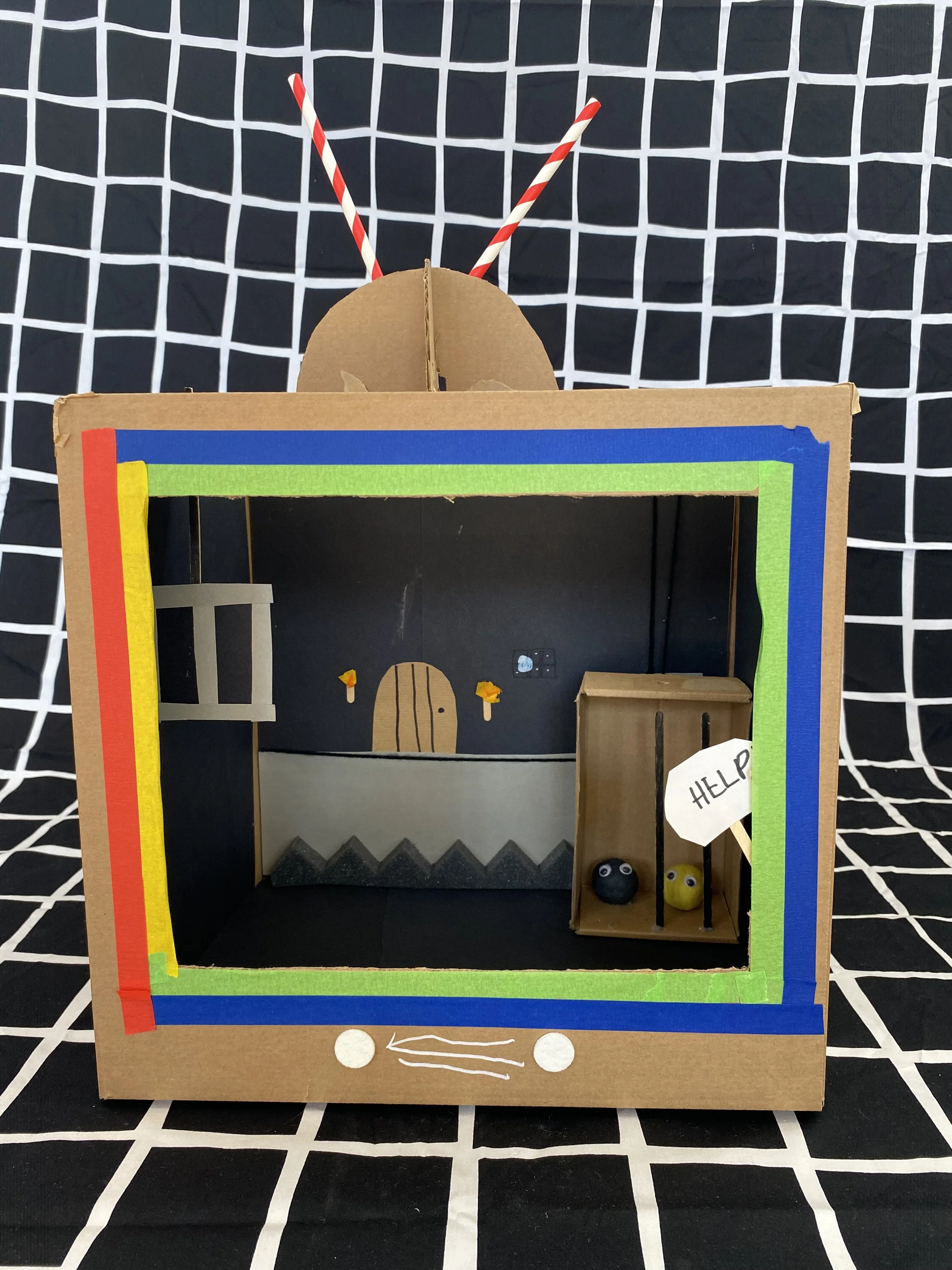 A cardboard puppet theater decorated with colorful tape and featuring a small door with a face and small decorations. Two black balls with googly eyes are inside the theater, with a small sign reading "Help" next to them. The background is a black and white grid pattern.