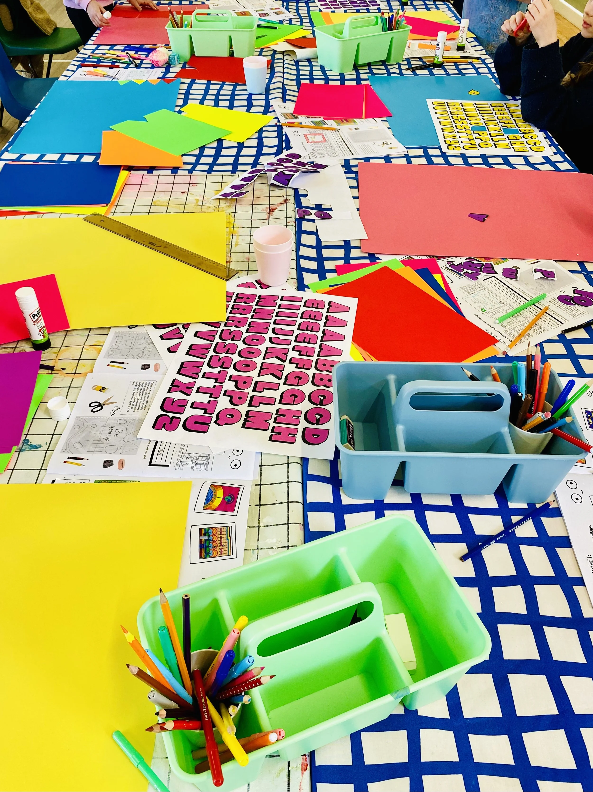 A colorful craft table with various sheets of colored paper, scissors, glue sticks, and pencils. There are green plastic bins holding markers and other supplies, along with printed sheets of alphabet letters, and a ruler. The table is covered with a blue and white checkered tablecloth, and some children's hands are visible working at the table.