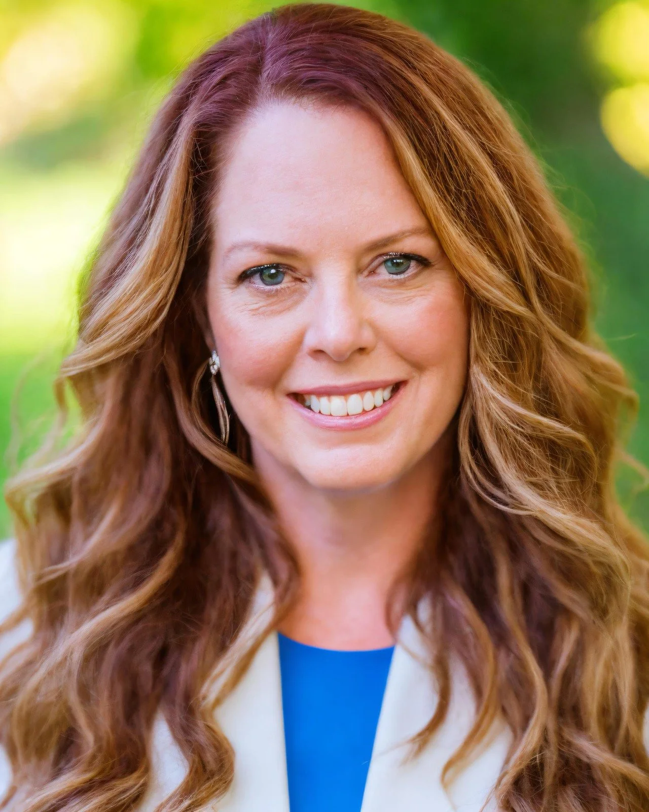 A close-up portrait of a smiling woman with long, wavy auburn hair, wearing a light blazer over a blue top, with a soft green outdoor background.