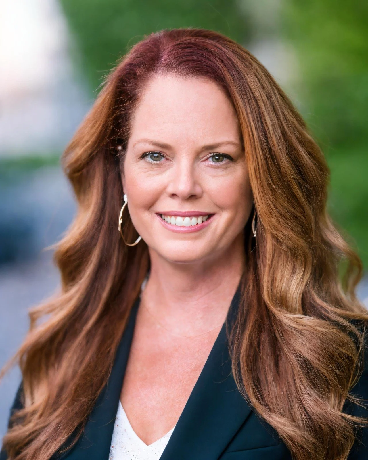 Professional outdoor headshot of a smiling woman with long, wavy auburn hair wearing a black blazer and gold hoop earrings with a blurred greenery background.