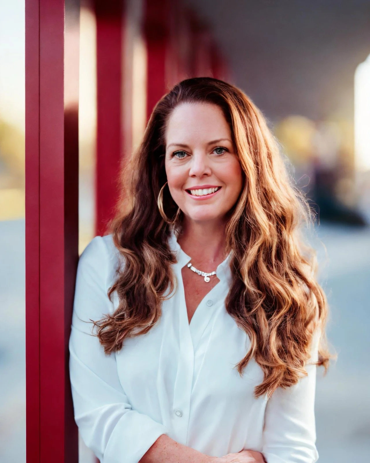 A portrait of a smiling woman with long, wavy auburn hair wearing a white blouse, standing outdoors beside a red wall with a softly blurred urban background.
