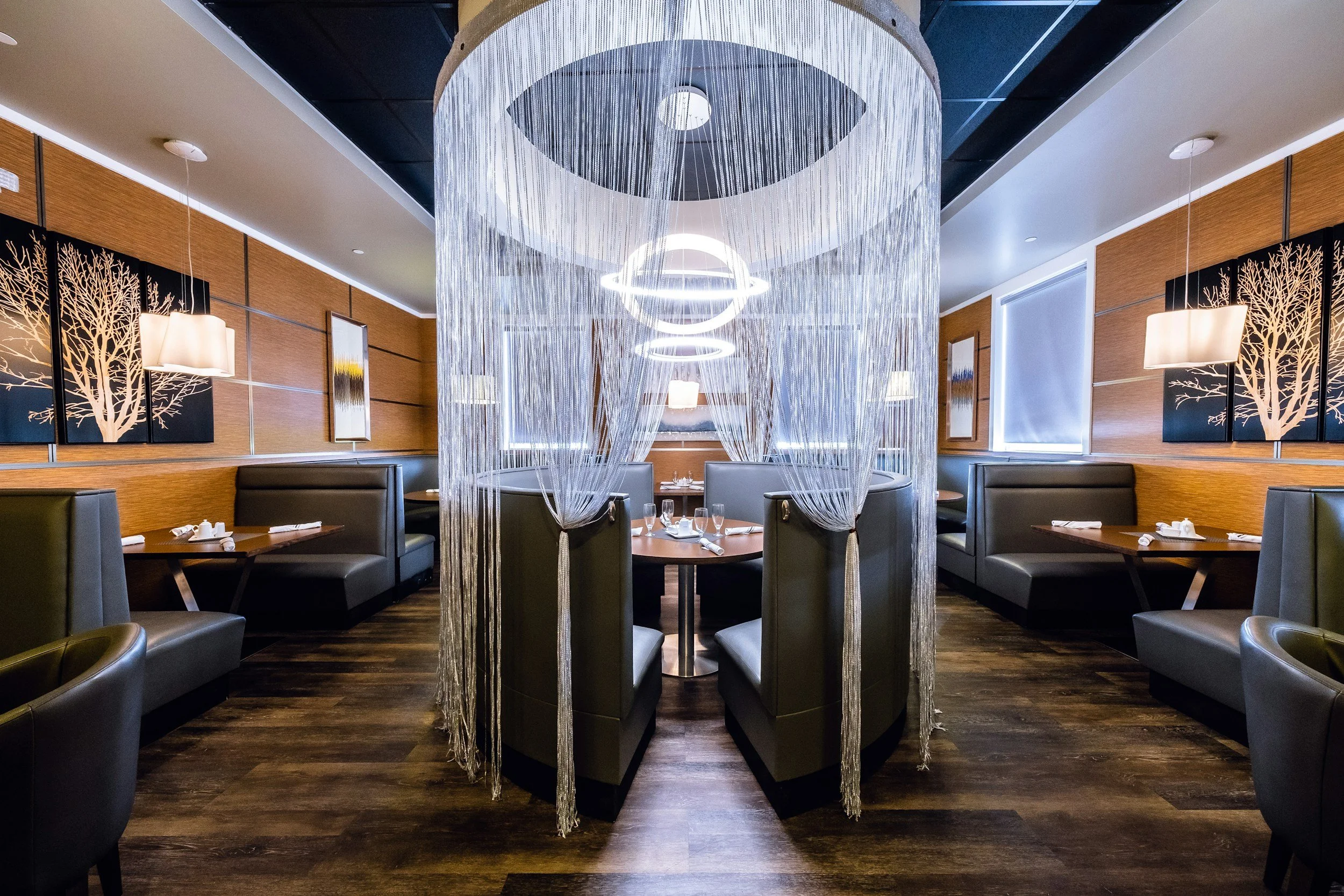 Salle à manger dans un restaurant élégant, avec des murs en bois, des lampes suspendues, des œuvres d'art représentant des arbres, et un éclairage moderne au plafond.