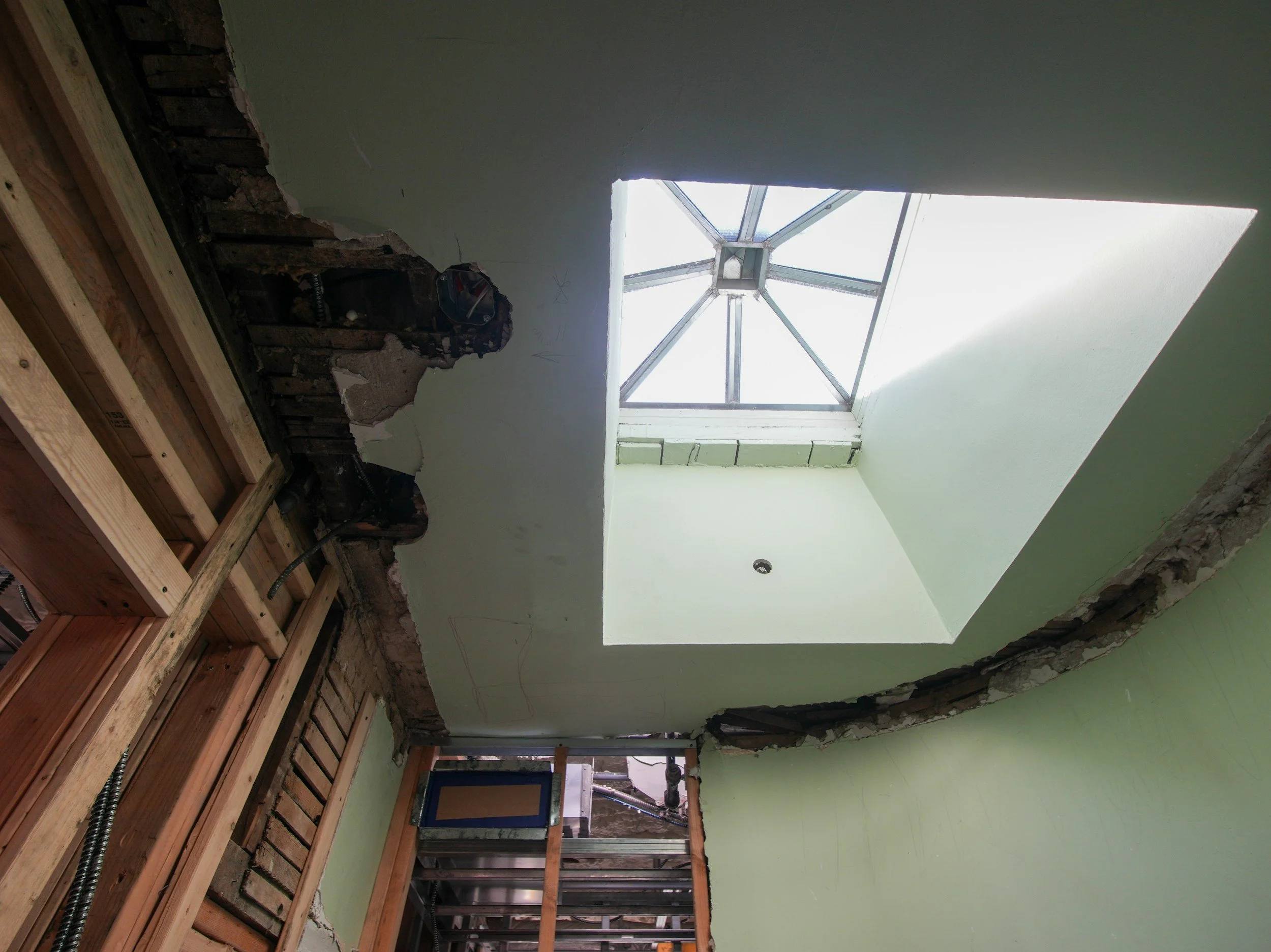 Travaux de rénovation intérieure avec une lucarne installée dans le plafond, murs en cours de démolition avec des matériaux endommagés, structure en bois visible, lumière naturelle entrant par la lucarne.