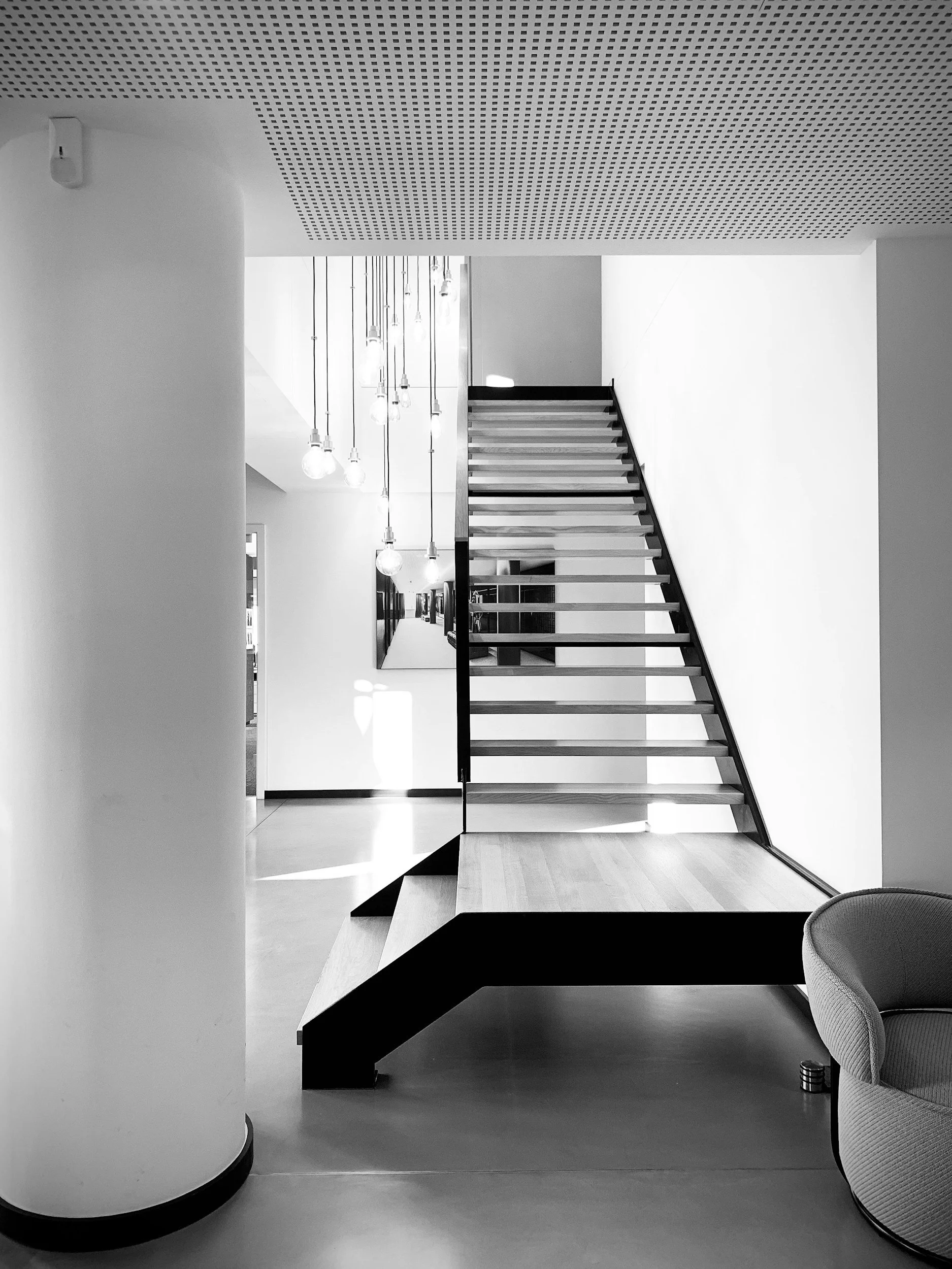 Interior moderne en noir et blanc avec un escalier en bois et métal, un plafond perforé, et des lampes suspendues, dans un espace résidentiel ou professionnel.
