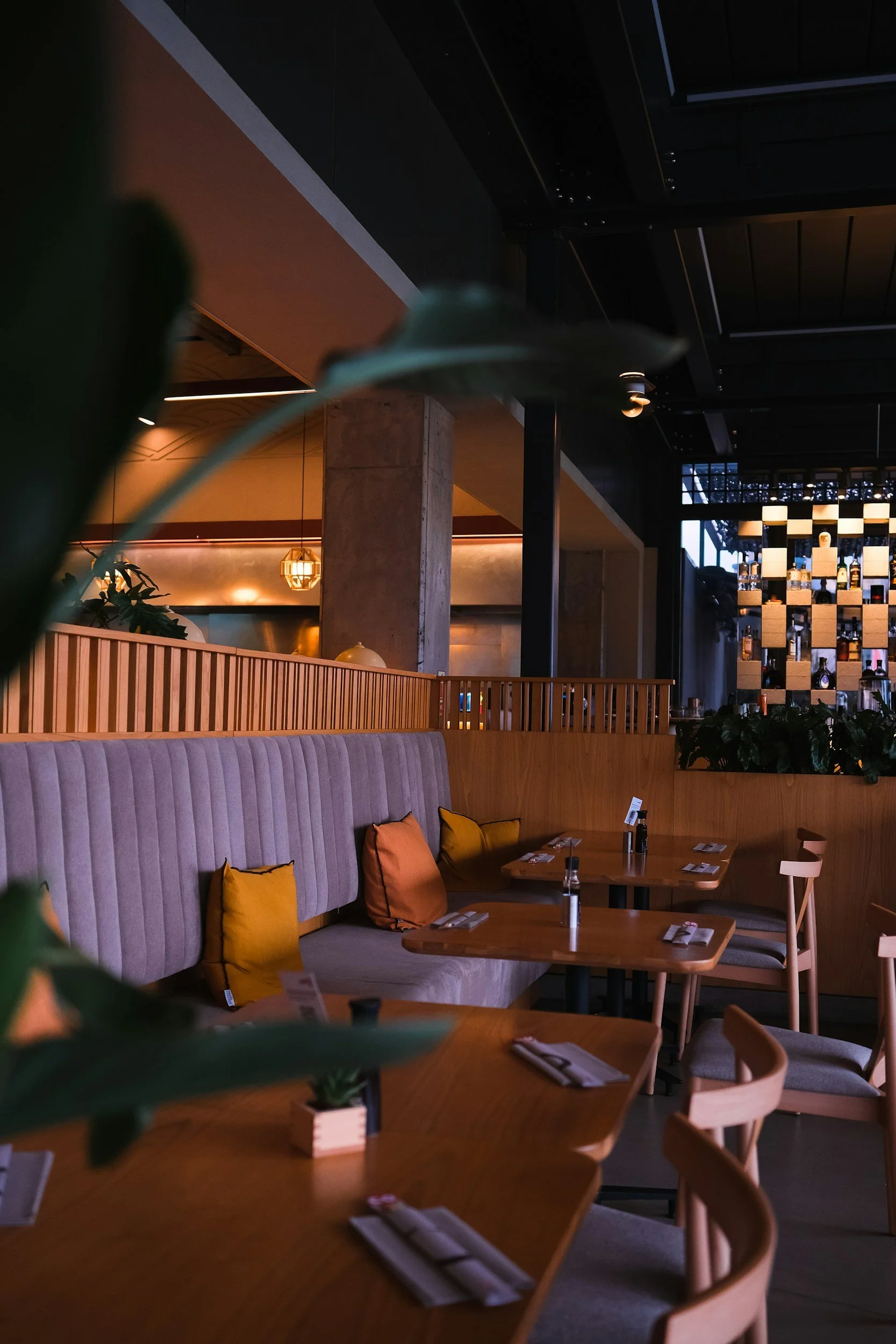 Intérieur d'un restaurant moderne avec des tables en bois, des coussins orange, une banquette rembourrée, des plantes vertes, une cloison en bois, une décoration chaleureuse, et un éclairage tamisé.