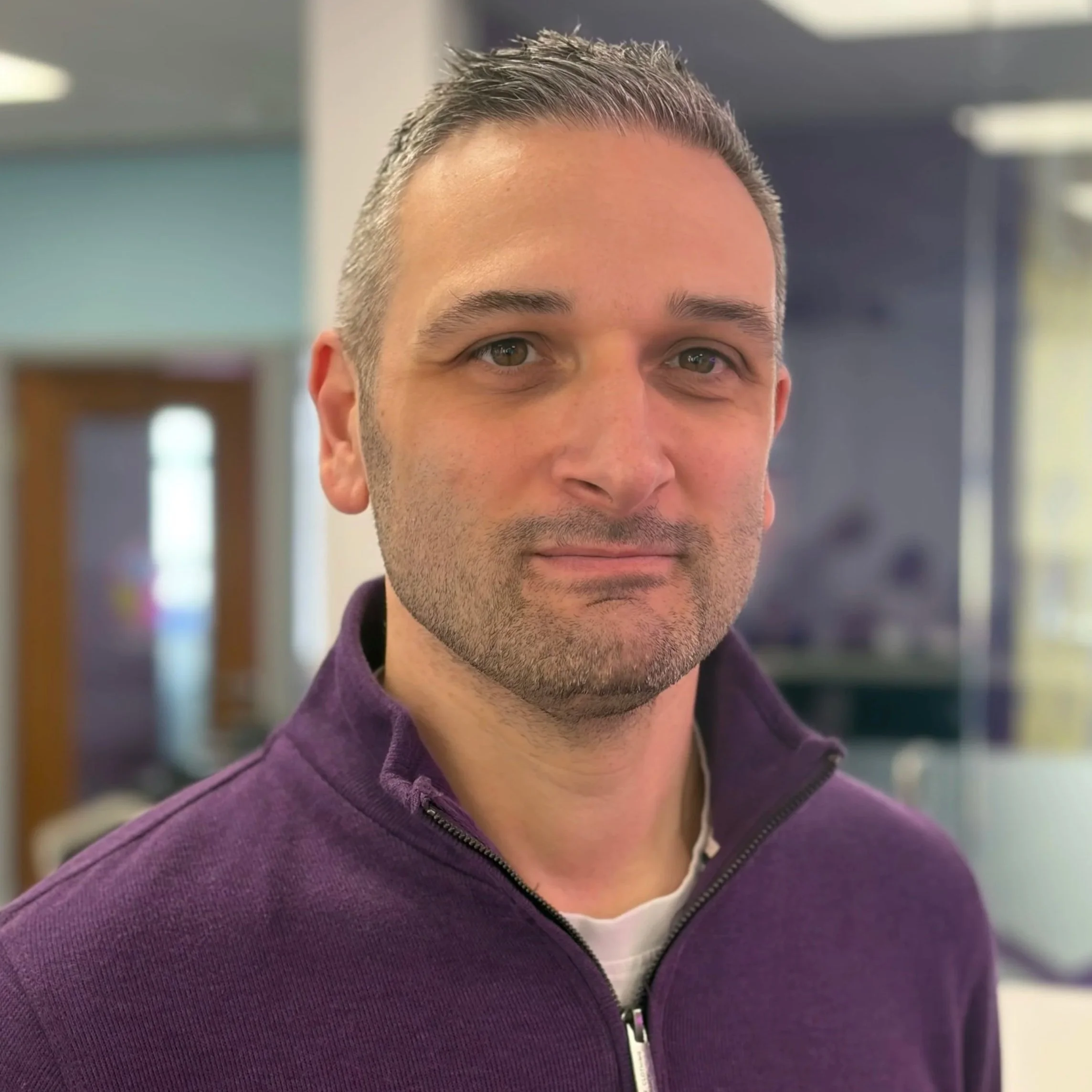A man with short dark hair, a beard, and light skin wearing a red collared polo shirt stands in front of a colorful, miniature cityscape background.