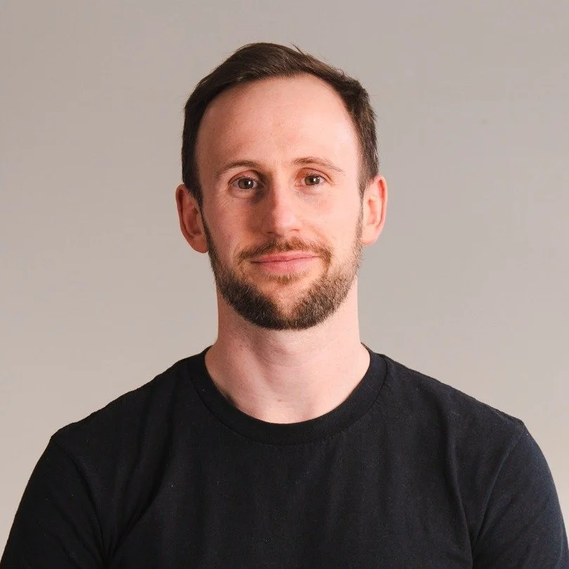 A man with short brown hair and a beard, wearing a black t-shirt, posing against a plain light gray background.