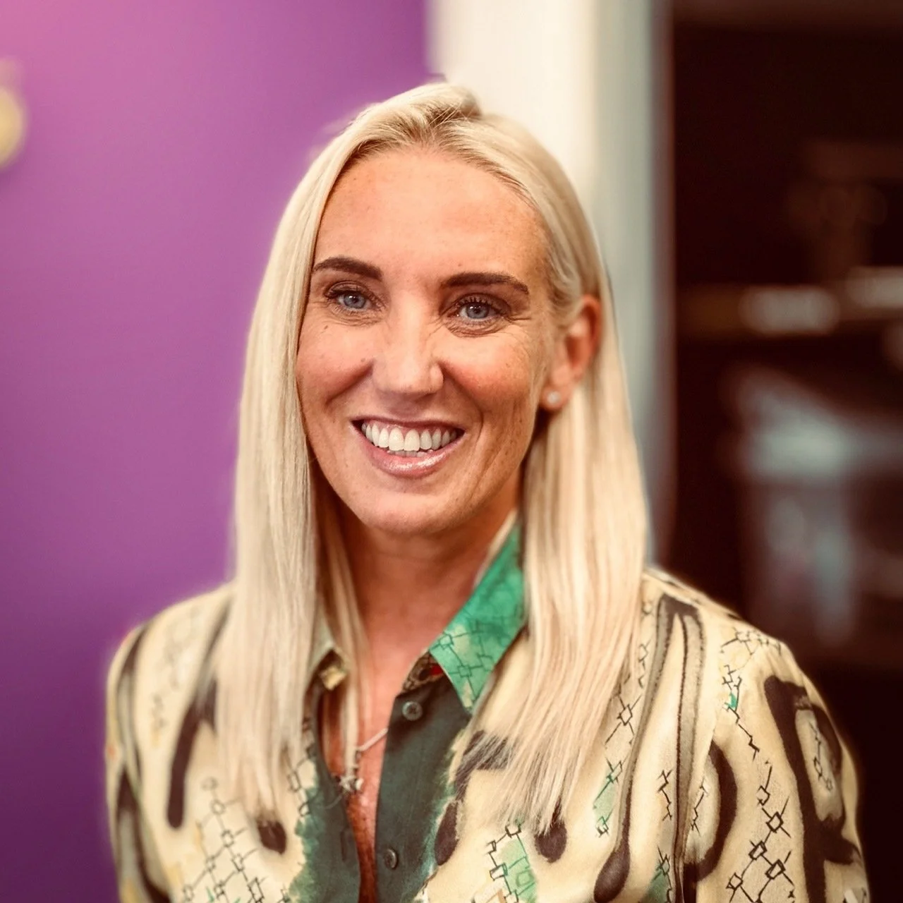A smiling blonde woman with blue eyes, wearing a patterned shirt with green accents, standing in front of a purple background.