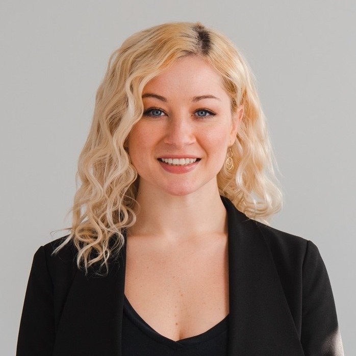 Portrait of a woman with blonde, curly hair, smiling, wearing a black blazer and earrings, against a plain background.