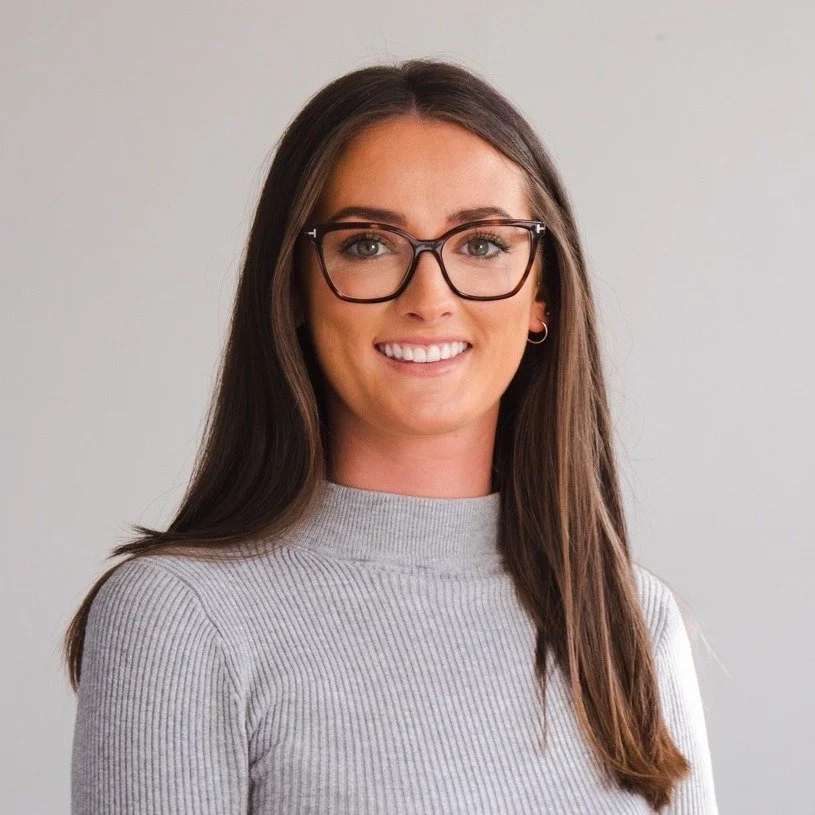 A woman with long brown hair, glasses, and a gray sweater smiling at the camera against a plain light background.